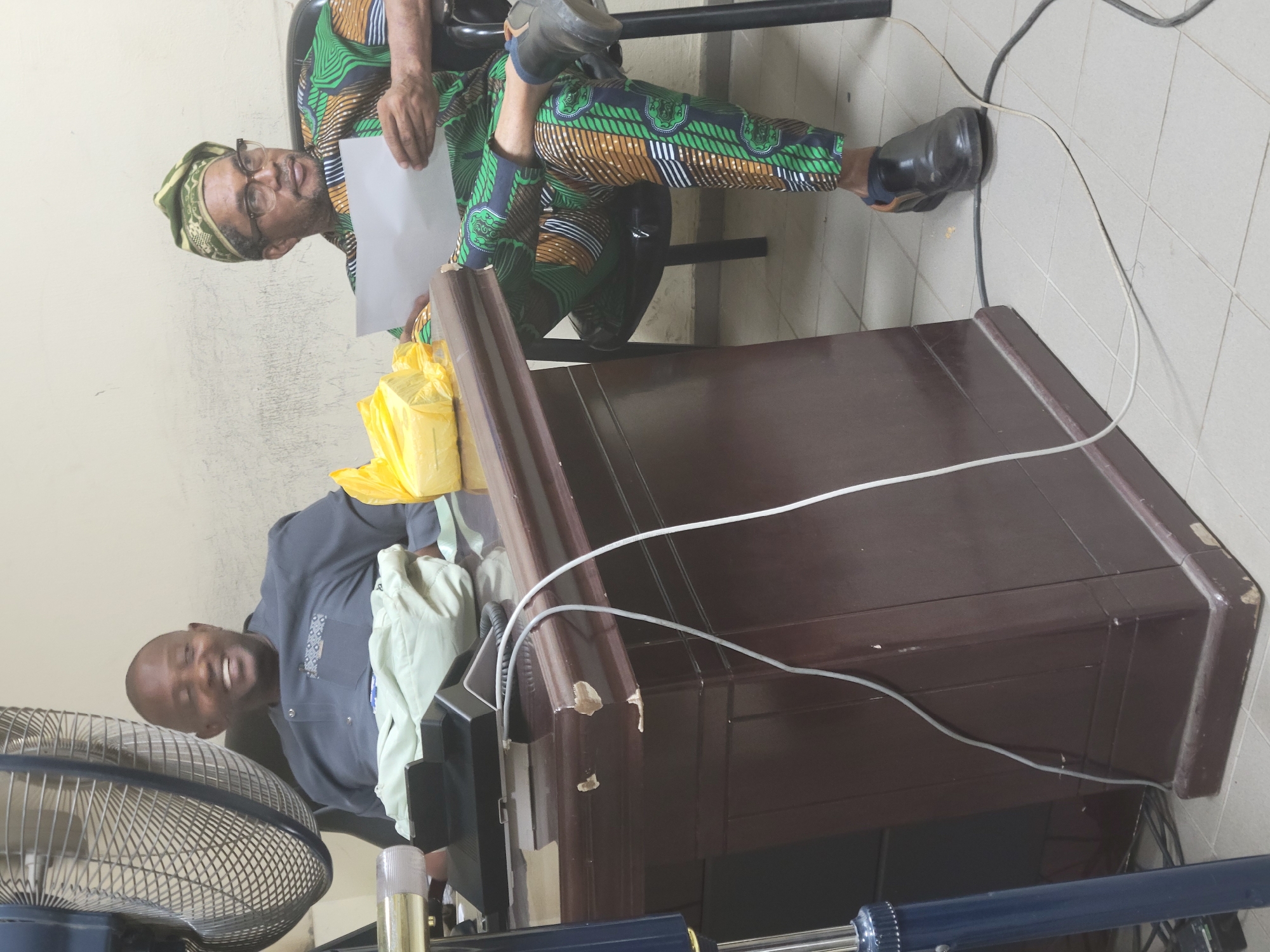 The image captures an indoor scene featuring two men in what appears to be an office or administrative setting, consistent with the Abuja, Nigeria context.
In the foreground, a bald, light-skinned man wearing a dark grey or blue traditional shirt with a patterned pocket is seated. He is smiling broadly and looking towards the viewer, appearing friendly and relaxed. A large metallic floor fan is positioned in front of him, its cage visible.
In the background and to the right, an older man with glasses is seated on a black chair. He is wearing a distinctive green, brown, black, and white patterned traditional outfit, likely made from Ankara fabric, with a matching headscarf. He holds a plain white sheet of paper in his hands and is also looking towards the viewer.
Between the two men is a dark brown wooden desk or console, which shows signs of wear with chipped edges. A yellow plastic bag is partially visible behind the desk, near the man in the patterned attire. Several white and dark electrical cables are visible, running across the top of the desk and along the tiled floor, suggesting connected equipment is present, though not fully visible. The walls are painted a light beige or cream color, and the floor is covered with light-colored tiles. The presence of the fan suggests a warm environment. No discernible text is visible in the image.