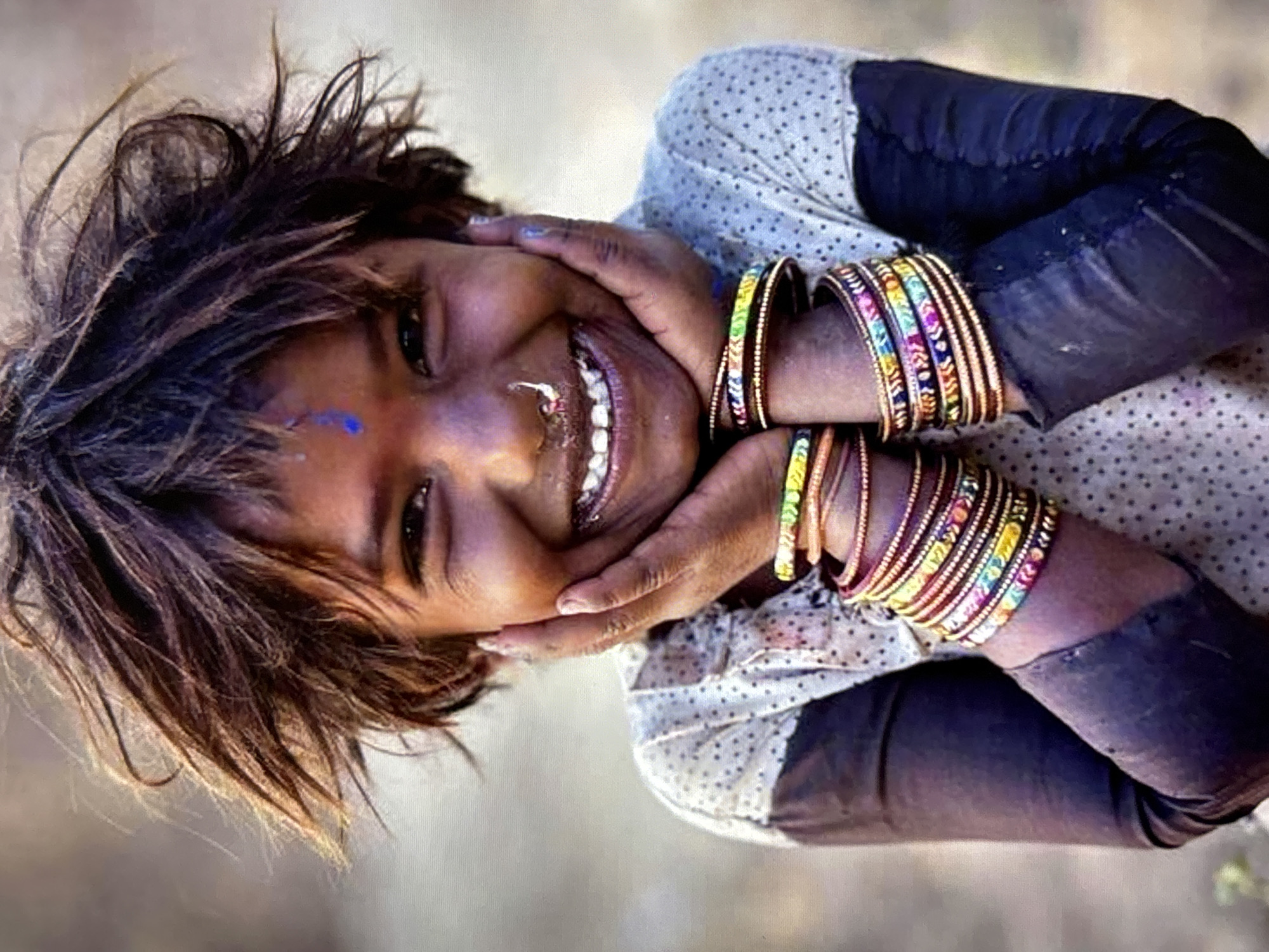 The image shows a close-up, sideways shot of a young person's face, which is tilted. Their hands are clasped to their cheeks, and their mouth is open in a wide smile, revealing teeth. A blue mark is visible on their forehead. They have dark, messy hair and are wearing a patterned shirt with dark sleeves. Multiple colorful bangles are stacked on their wrists. The background is blurred and appears to be an outdoor environment with muted, earthy tones, suggesting it is a natural or unpaved setting. There is no indication of the city of Orihuela, Spain, in the image.