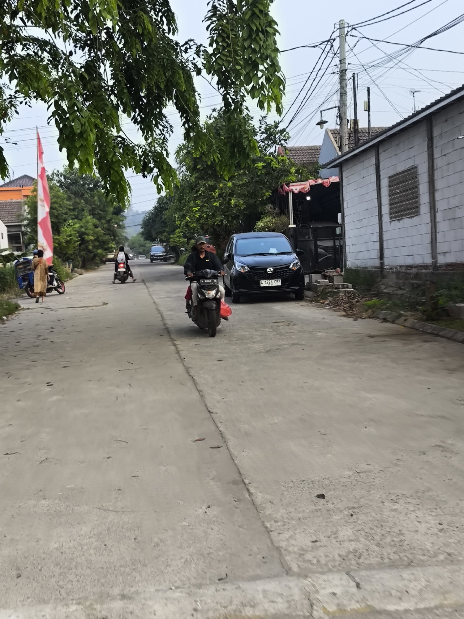 In Cilegon, Indonesia, on a cloudy day, a street scene unfolds. A person rides a scooter in the foreground, wearing a black jacket and a red bag hangs from the back of the scooter. To the left, another person walks away from a parked three-wheeled motorcycle, wearing a beige dress. Further down the street, a person on a scooter wearing a white backpack travels away from the viewer. A black car, a Daihatsu, with license plate L 1724 CBP, is parked on the right side of the road. Buildings line the street, including a white brick structure with a patterned window. Trees and electrical poles with numerous wires are visible. A red and white Indonesian flag flutters in the breeze. The road is paved and appears to be in a residential or semi-rural area.
