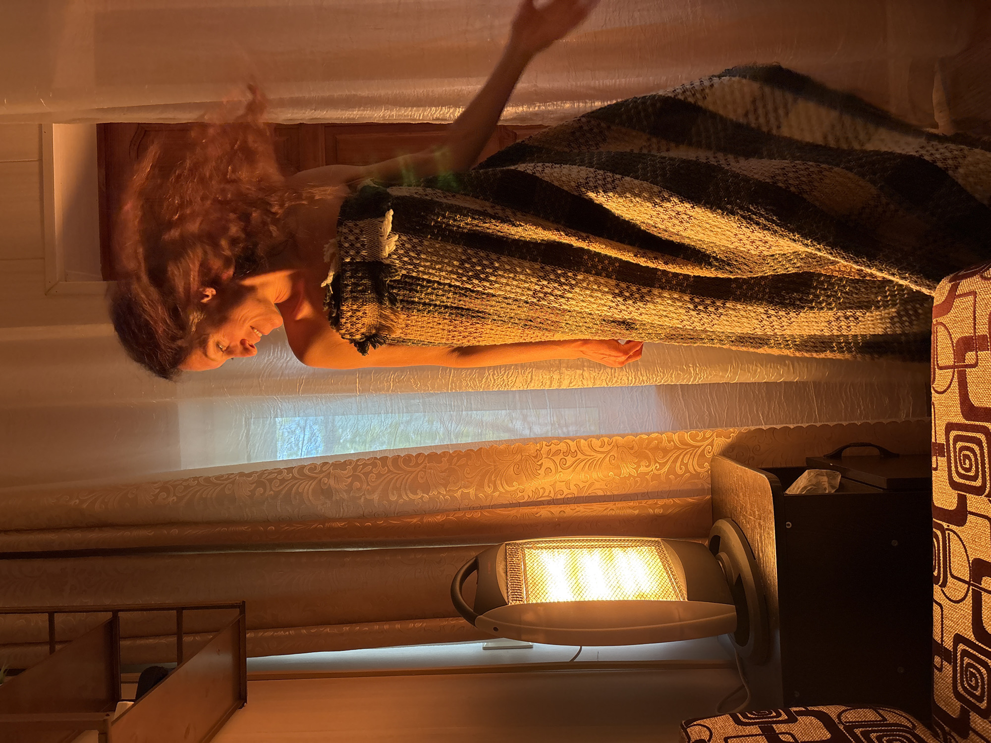 A woman with long, reddish-brown hair is lying under a brown and beige striped blanket in a room. Her head is near a wooden door frame, and she is smiling. One arm is outstretched above her head. She appears to be indoors, as suggested by the presence of curtains, a portable electric heater, and a couch. The scene appears to be at night or in a dimly lit space, accentuated by the warm glow of the heater. Location information is not explicitly available in the provided image.
