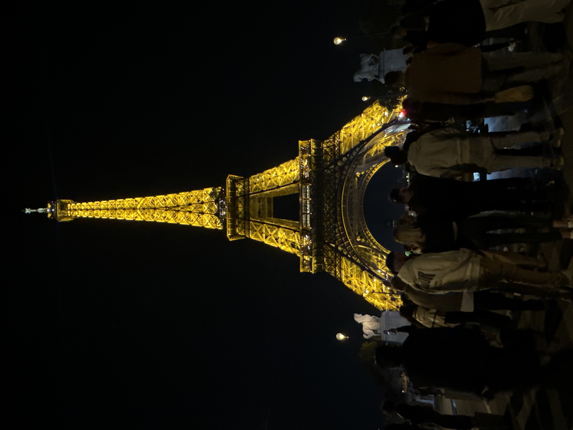 The image shows the Eiffel Tower at night, illuminated with yellow lights. A crowd of people stands near the base of the tower. Streetlights are visible. The location is Paris, France.