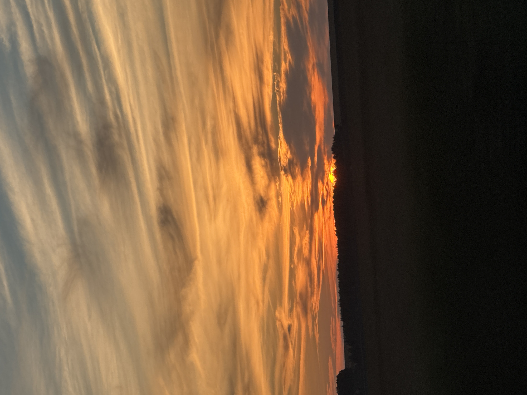 The image depicts a rural landscape at sunset. The sky is filled with dramatic clouds, painted in hues of orange, yellow, and subtle blue. The sun is low on the horizon, casting a warm glow. In the foreground, a dark silhouette of trees and rolling hills separates the sky from the land. The lower portion of the image is dominated by a dark, featureless field, suggesting nighttime is approaching. The scene is devoid of people and any specific structures, indicating a natural, undeveloped environment. The location is identified as Bol'shoe Aksakovo, Russia.