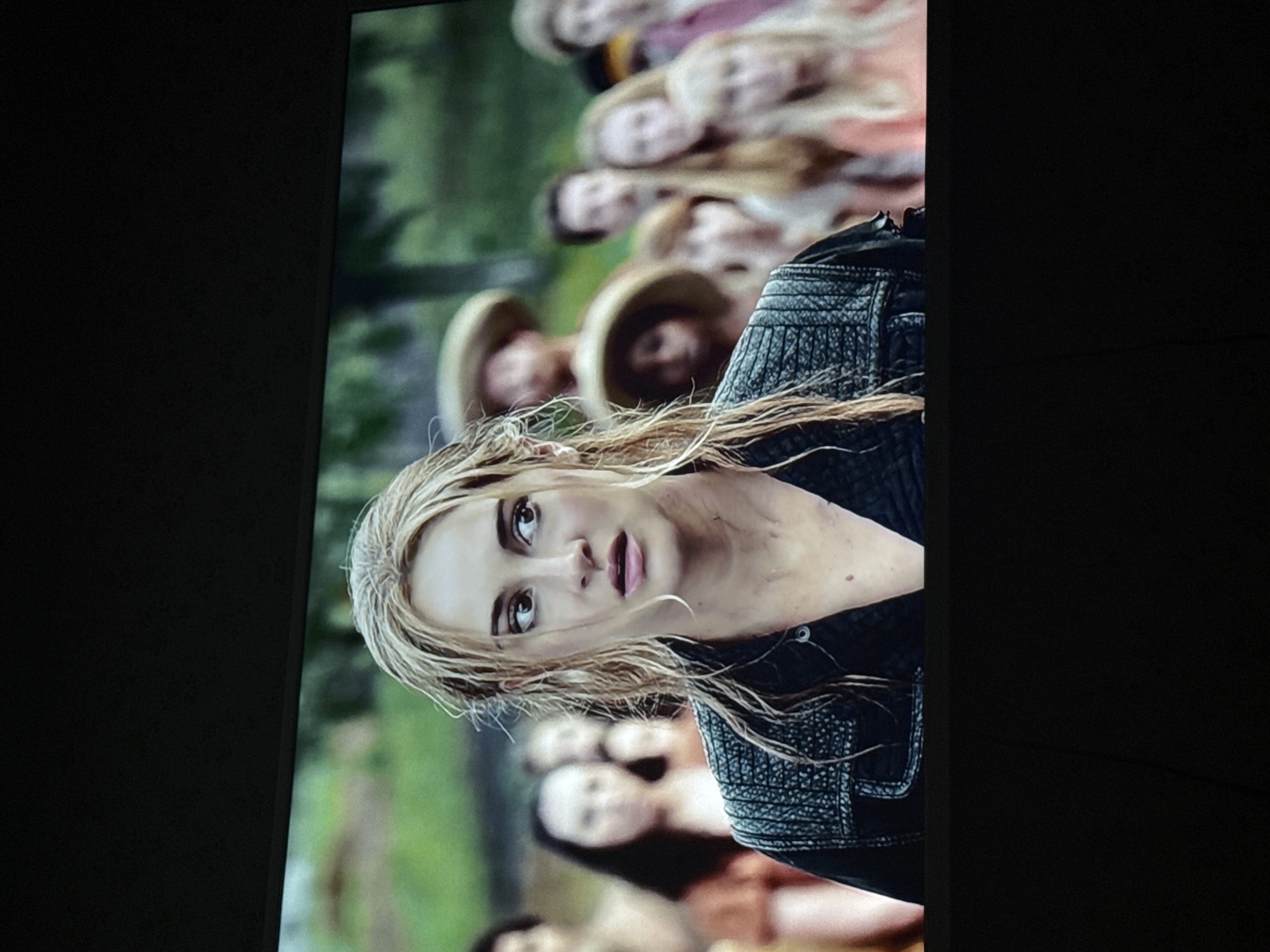 The image shows a scene displayed on a screen, likely a television. The scene depicts a fair-skinned woman with blonde, slightly wet hair, looking towards the viewer with a somewhat concerned expression. She is wearing a dark-colored, textured jacket. In the background, there is a blurry crowd of people; some are wearing straw hats, while others appear to have light-colored hair. The background is somewhat green, suggesting an outdoor, possibly wooded, environment. The location is not identifiable as Mytishchi, Russia or any specific location based on the given visual information.