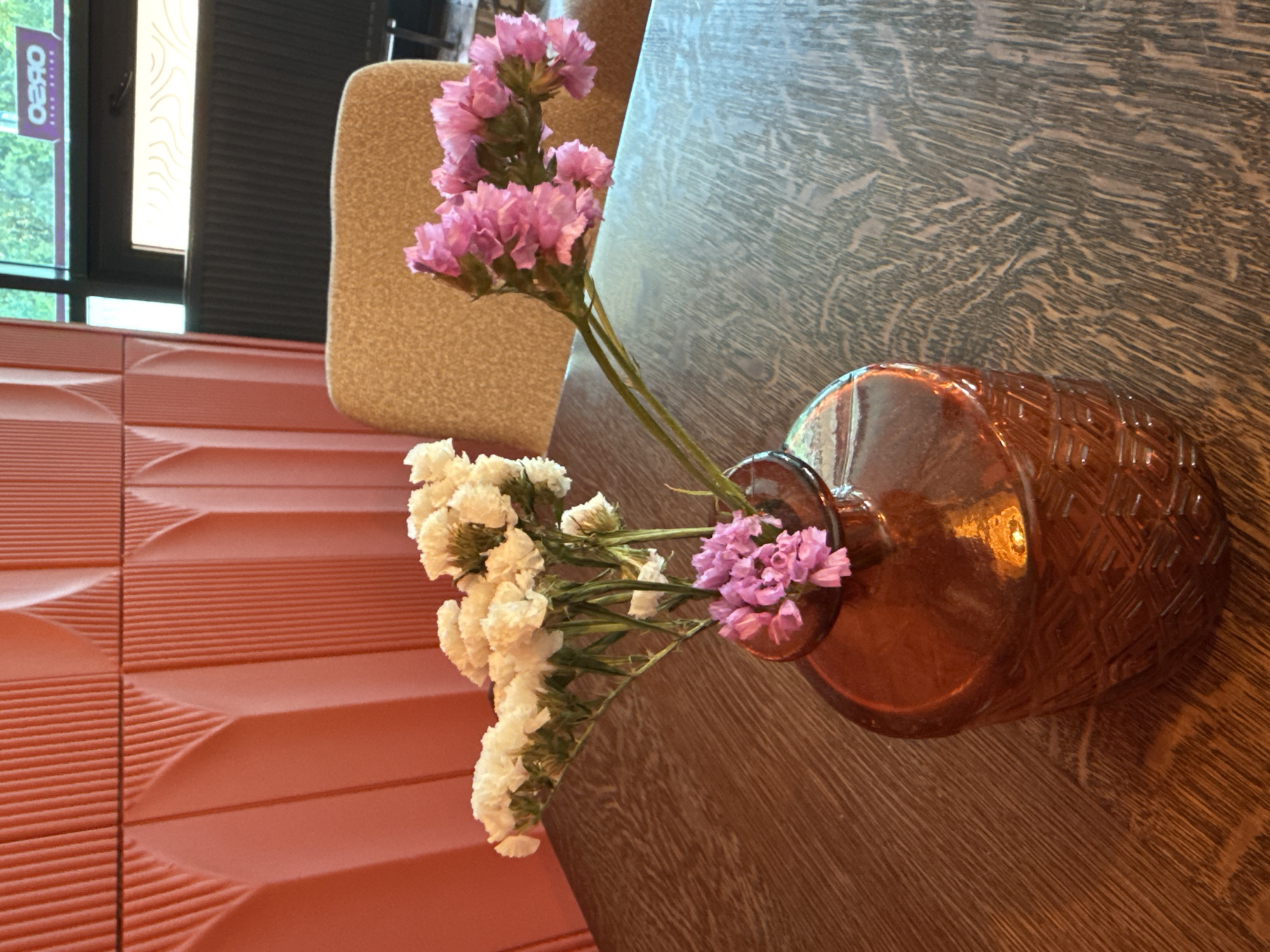 In Bălți, Moldova, an amber glass vase containing a bouquet of dried flowers sits on a wooden table. The bouquet is composed of white and pink flowers. In the background, a coral-colored wall with a ribbed texture is visible, along with a beige chair and a window with a view of greenery and a sign. The setting appears to be an indoor establishment, possibly a cafe or restaurant.