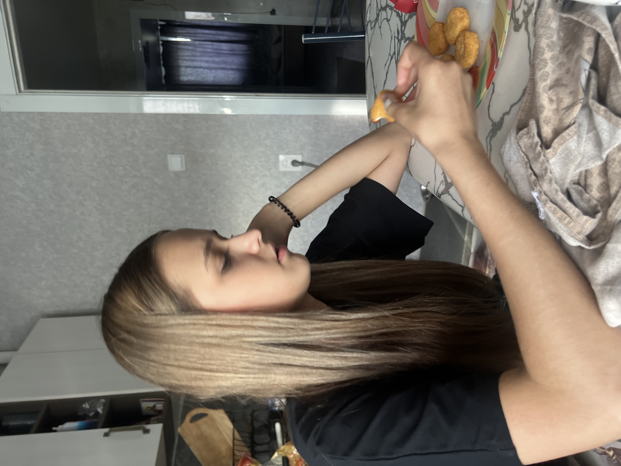 A fair-skinned person is eating from a plate of approximately four chicken nuggets. The person is wearing a black shirt and has long blond hair. The person is seated at a white countertop. A tan dishcloth is visible on the countertop. A section of a white wall is visible in the background. The scene is indoors in an unknown city.