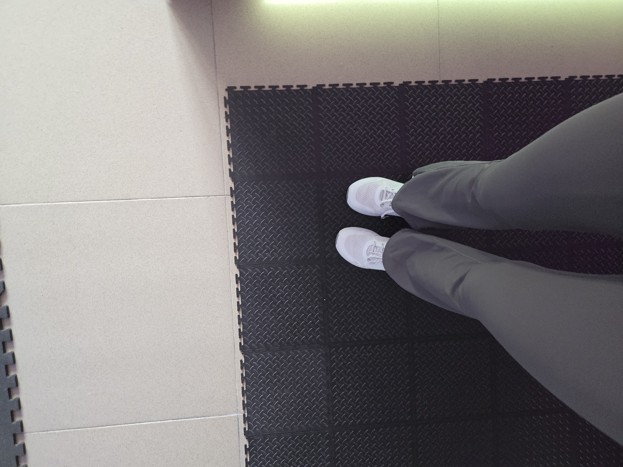 A first-person perspective shot shows a person's legs and feet, standing on black interlocking floor mats. The person is wearing grey pants and white athletic shoes. The mats are laid out in a diamond plate pattern and are bordered by light-colored floor tiles on the top and right side of the image. The floor tiles are a uniform light beige color with subtle speckling. The edge of the mats forms a jigsaw-like pattern where they connect. The lighting suggests an indoor environment with a bright, diffused light source coming from the right side of the frame, casting a soft shadow. The scene is devoid of any indicators of specific location within Krasnodar, Russia.