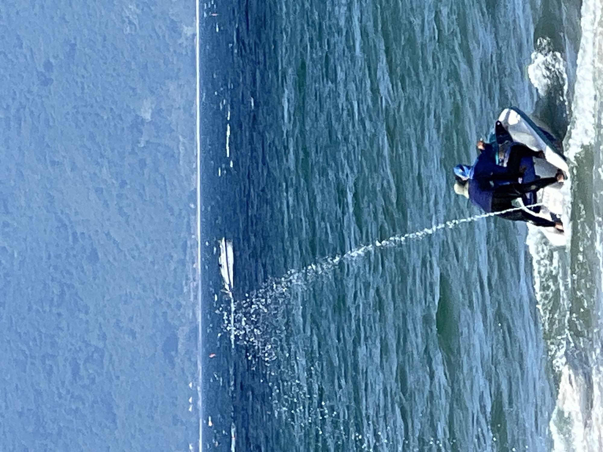 Two individuals, one wearing a blue hooded jacket and the other a beige hat and blue jacket, are riding a white and blue jet ski in the ocean. They are both facing away from the camera, with the person in the blue hooded jacket in front and the person in the beige hat behind. The jet ski is creating a wake and spraying water into the air. In the background, a white boat is visible on the water, and a large, forested mountain range can be seen in the distance. This scene is consistent with activities found at beaches along the coastline of Da Nang, Vietnam, such as My Khe Beach or Non Nuoc Beach.