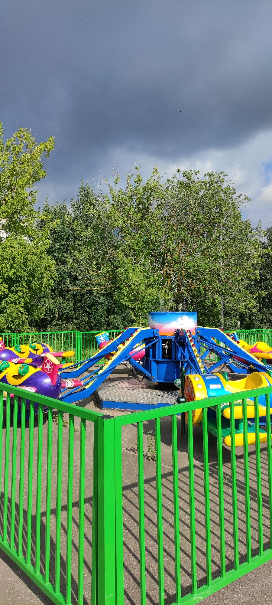 The image shows a children's amusement park ride in an outdoor setting. The ride features several colorful airplane-shaped cars mounted on arms that extend from a central blue platform. The cars are primarily purple and yellow with red accents and star designs. The ride is surrounded by a bright green metal fence. In the background, there are numerous green trees and a cloudy sky. The ground appears to be paved with concrete. The location is identified as Khimki, Russia. There are no people visible in the image.