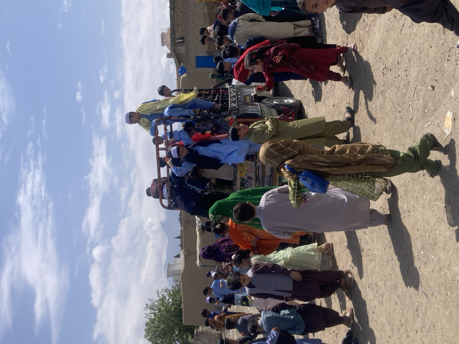 The image depicts an outdoor scene in Quetta City Tehsil, Pakistan, under a partially cloudy blue sky. A large group of individuals, primarily children and young adults, are gathered around a gray utility truck with an open bed. The truck's bed and metal frame are occupied by numerous young males in traditional shalwar kameez, with one individual in a light blue outfit and yellow scarf standing prominently on the truck's side rail. Others are seen actively boarding or disembarking.
On the unpaved, dusty ground surrounding the vehicle, more people are congregated. A girl in a green shalwar kameez and a brown patterned scarf, carrying a blue shoulder bag, walks away from the camera in the foreground. Another girl in a dark red outfit is seen gesturing or interacting with an unseen individual near the truck. Numerous other children and adults, including women in colorful traditional dresses and head coverings, are present. Shadows cast by the individuals and objects indicate bright daylight.
In the background, single-story, light-colored buildings with flat roofs are visible. Distant hazy mountains rise along the horizon, and a distinct white structure, possibly a mosque or significant building, is visible to the left. A single green tree is also present on the far left. Small pieces of white litter are scattered on the ground.