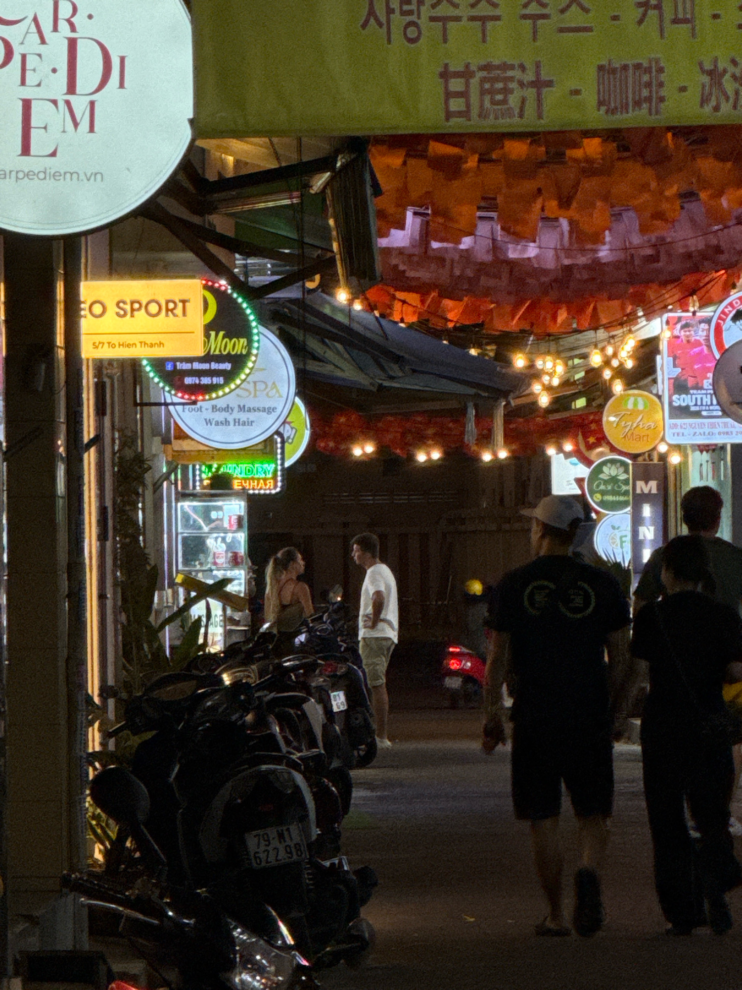 The image captures a street scene at night in Nha Trang, Vietnam, likely on a pedestrian walkway or a side street with shops and businesses. The overall atmosphere is illuminated by neon signs, string lights, and the warm glow from shop interiors.
In the foreground, several motorcycles are parked closely together, indicating a common mode of transportation in the area. One license plate is partially visible, reading "79-M1 622.98".
Several individuals are present, blurred due to motion or low light. On the left, a couple stands near a doorway, with the woman facing left and the man facing towards her. Further to the right, a man in a light-colored shirt and shorts stands looking towards the left. In the center-right, two figures, possibly men, are walking away from the camera. One is wearing a black t-shirt with a circular logo and dark shorts, while the other is wearing black pants and a dark top.
Various signs are visible, advertising businesses. To the left, a large circular sign with the text "CARPE DIEM" and "carpediem.vn" is prominent. Below it, a rectangular sign reads "EO SPORT" with an address "5/7 To Hien Thanh". Adjacent to this, a circular neon sign displays "Moon" and below that, "Trâm Moon Beauty" and a phone number. Another sign indicates "SPA" and services like "Foot - Body Massage" and "Wash Hair". Further down the street, other illuminated signs for "Tyfha Mart" and "Oasis Spa M" are visible.
Overhead, red and orange decorative banners or lanterns are hung, creating a festive or commercial display. The ground appears to be asphalt or concrete. The background shows the interiors of shops with shelving and products, as well as more lighting, suggesting a busy commercial district. The image captures the vibrant nightlife of Nha Trang, with a mix of local life and commercial activity.