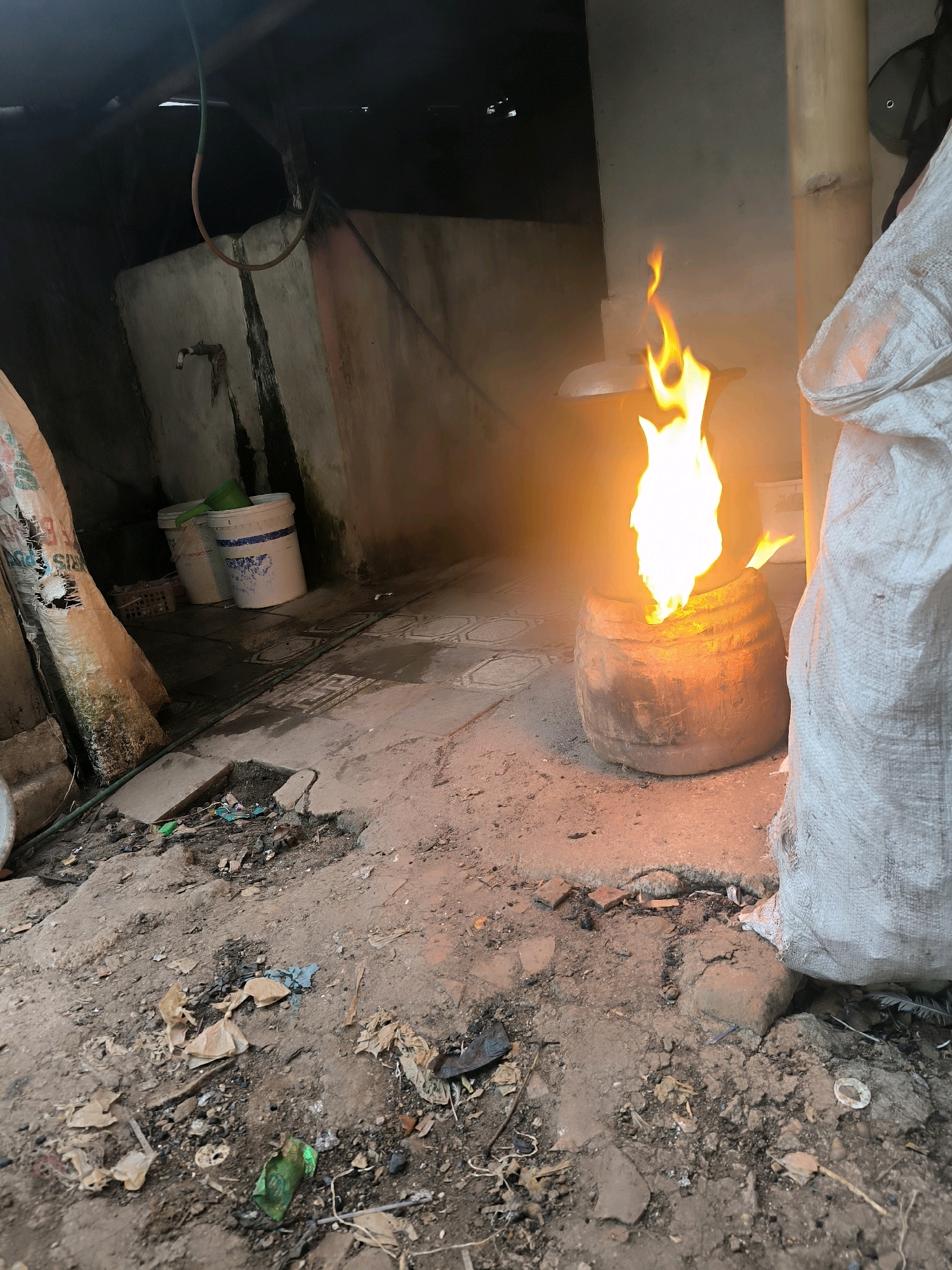 In a dimly lit outdoor area, likely in Cilengkrang, Indonesia, a fire burns intensely within a large, cylindrical pot or furnace. The fire, a vibrant orange and yellow, casts a strong glow on its immediate surroundings. To the right of the fire, a large, white, coarse-woven sack is partially visible, leaning against a rough, vertical structure that appears to be made of bamboo. The ground is uneven and covered with dirt, debris, and scattered litter, including plastic fragments and dried leaves. In the background, a concrete wall with some staining and weathering is visible, along with a metal faucet. Two white buckets, one with a green lid, are placed near the wall. A coiled hose is also present, hanging from above. The overall environment suggests a utilitarian or perhaps somewhat neglected outdoor space. There are no people visible in the image.