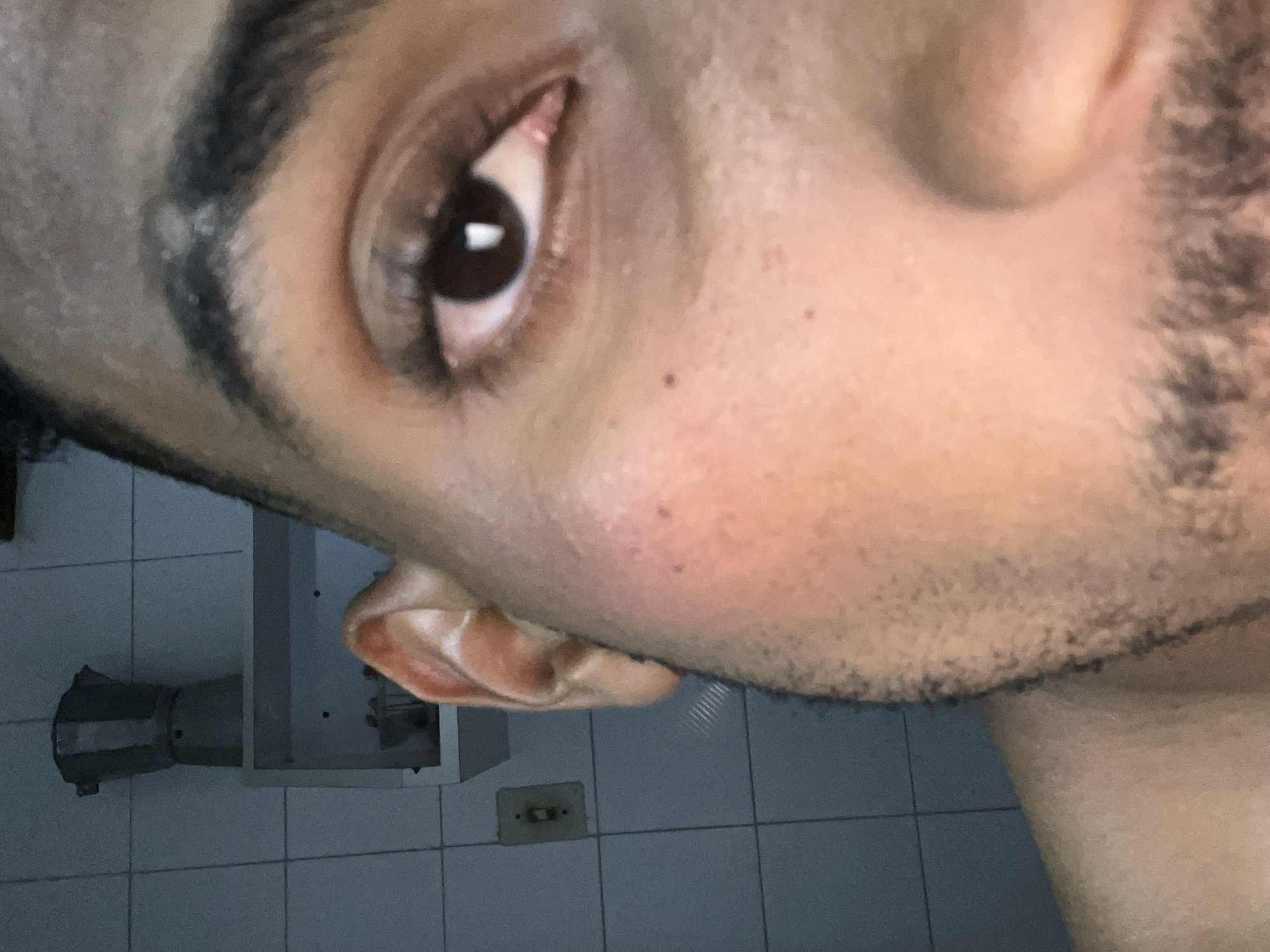 The image is a close-up, unposed, medium-shot photograph of the right side of a person's face, including their eye, cheek, ear, and a portion of their neck. The person has brown skin and dark eyebrows. A small dark mole is visible on the cheekbone below the eye. The background consists of tiled walls and a shelf with what appears to be a moka pot. The scene is indoors, likely in a domestic setting such as a kitchen. The lighting is dim, creating shadows on the face and reducing the clarity of the background details. There are no other people or animals present in the image. The photograph is taken from a slightly low angle, looking up at the subject. The overall impression is of an informal self-portrait or snapshot. The location in Porlamar, Venezuela, cannot be determined from the visual information in the image.