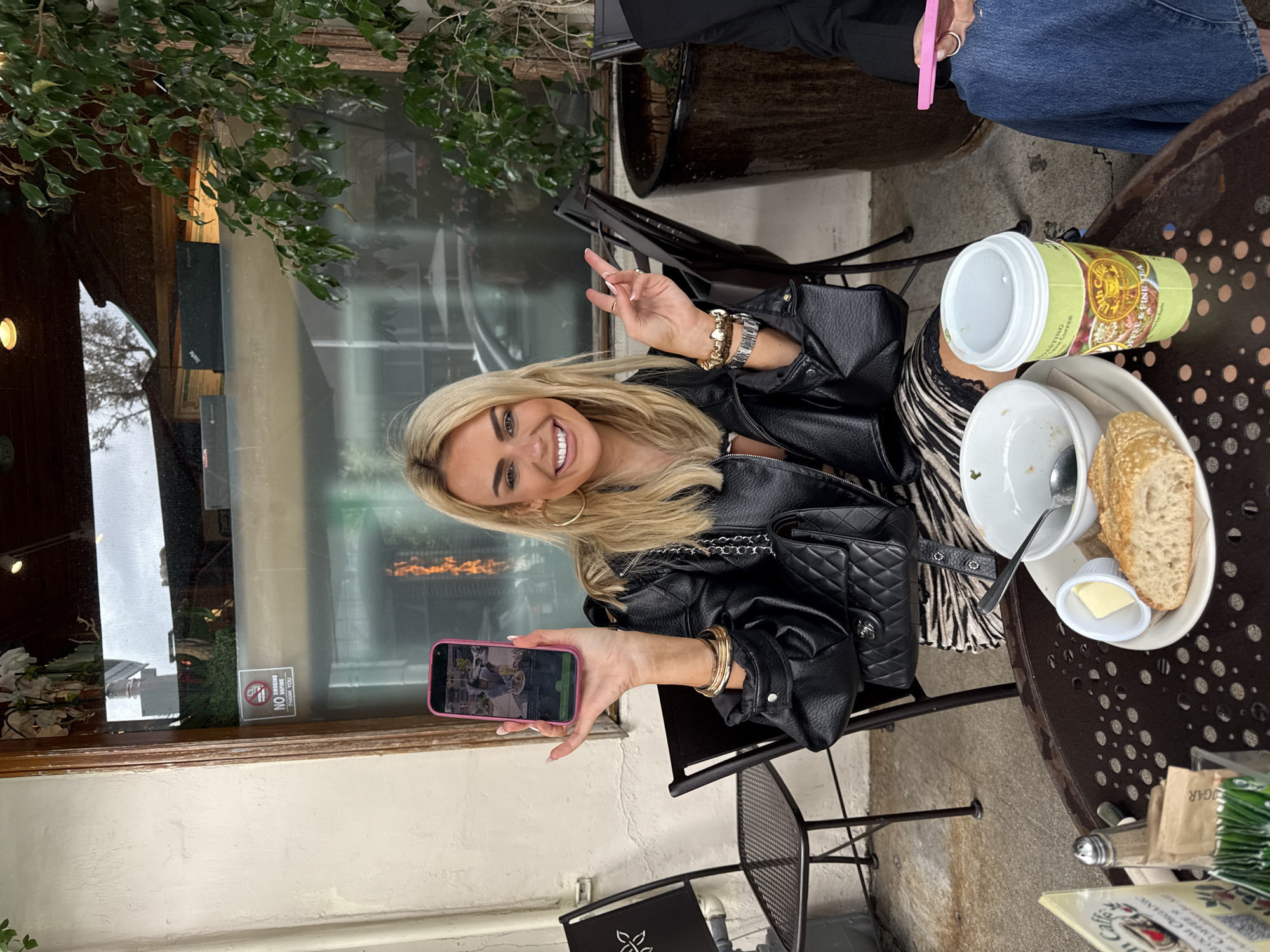 Here's a detailed description of the scene in the image:
**Setting:**
The image appears to be taken at an outdoor cafe or restaurant in West Hollywood, California. This is deduced from the "Urth Caffe" cup and the "No Smoking/Vaping" sign, which is identifiable as belonging to Urth Caffe and mentioning the city of West Hollywood.
**People:**
The primary subject is a blonde woman who is smiling and holding up a smartphone. She is wearing a black leather jacket, jewelry (including a watch and bracelets), and is making a peace sign with one hand. Another person wearing blue jeans is partially visible in the upper right corner, suggesting she is sitting with someone.
**Objects:**
* **Table setting:** A plate with the remains of food (likely soup and bread), a small container of butter, a coffee cup from "Urth Caffe," a salt shaker, sugar packets, and a menu are on the table.
* **Furniture:** Wrought iron chairs and a table are visible.
* **Background:** A glass window, greenery, and hints of building exteriors are visible in the background, typical of a storefront cafe.
* **Accessories:** A quilted black handbag is resting near the plate.
**Actions/Interactions:**
The woman is holding her phone, seemingly showing an image or taking a photo, possibly of the meal or setting. Her smiling expression suggests she's enjoying the experience.