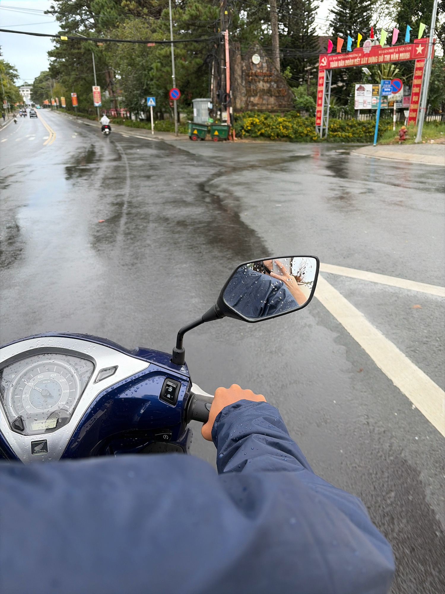 The image captures a first-person perspective of riding a motorcycle on a wet, paved road in Da Lat, Vietnam. The road is reflective due to the rain, showing streaks and puddles. Trees line the left side of the road, and a building with a white facade is visible in the distance. On the right side of the road, there is a red archway with Vietnamese text and colorful flags. A sign with Vietnamese text and a red circle with a horizontal bar is also present, indicating a restriction. In the background, there is a large stone monument with inscriptions.
In the foreground, the handlebars and part of the dashboard of a blue and silver motorcycle are visible. The dashboard has a speedometer with markings up to 140 km/h and several indicator lights. A person's right hand, wearing a blue jacket with raindrops on it, is gripping the right handlebar. A side-view mirror on the motorcycle reflects another person, also wearing a blue jacket, holding a smartphone to take a selfie. The reflected hand shows a light skin tone and is holding the phone in a horizontal position. The background in the mirror shows trees and the sky.
Several other vehicles are present on the road, including a white car and a motorcycle with a person wearing a white umbrella-like covering. The overall scene depicts a rainy day commute or travel on a city street.