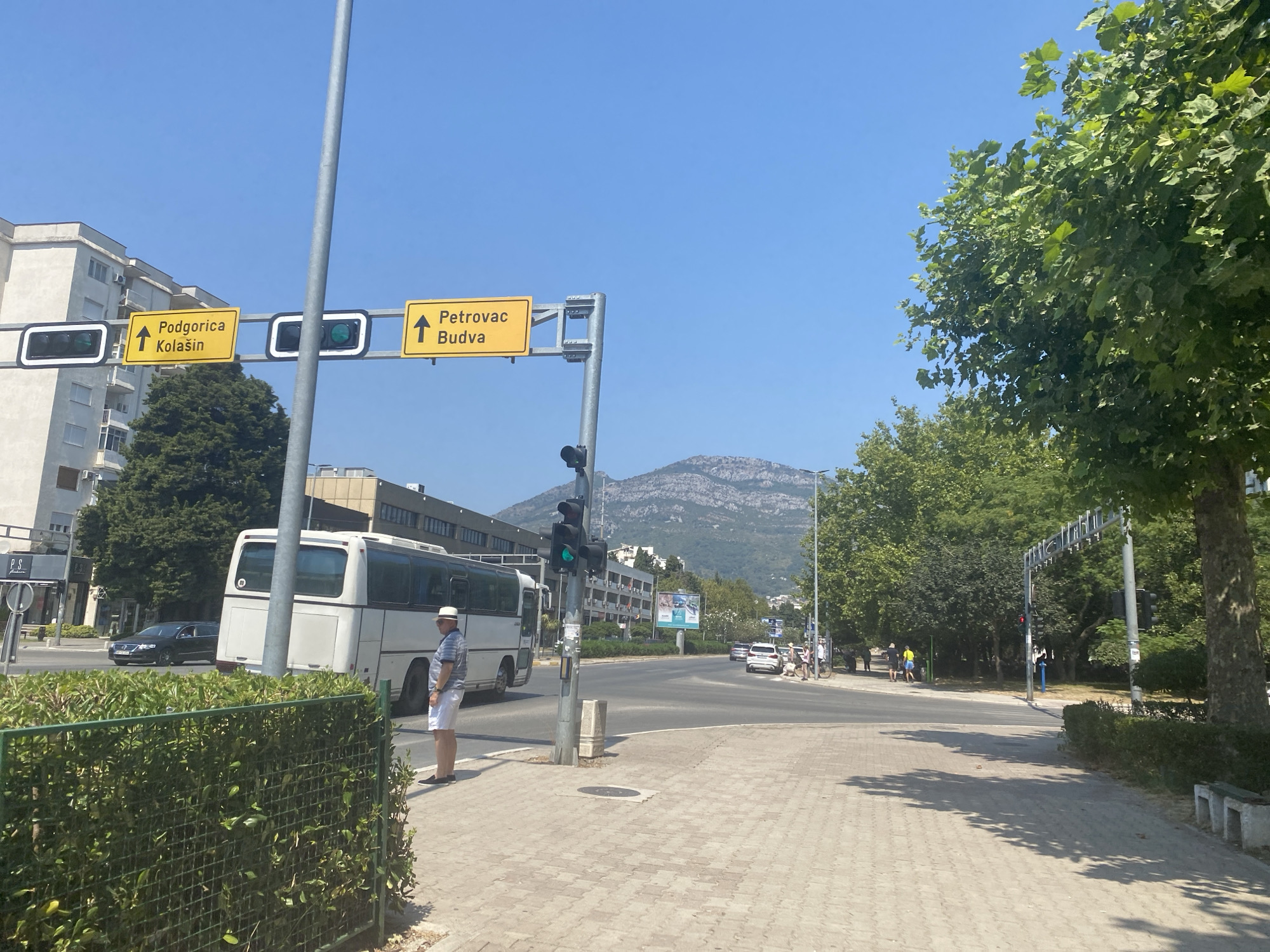 This is a wide shot of a street in Bar, Montenegro on a sunny day. In the foreground, a paved walkway runs alongside a neatly trimmed hedge. A traffic light hangs overhead, showing a green light, and two yellow directional signs with black lettering are mounted on the same pole. The signs indicate "Podgorica Kolašin" and "Petrovac Budva" with upward arrows. To the left, a white bus is parked on the side of the road, with a man in a striped shirt and white shorts standing on the sidewalk next to it. Beyond the bus, a black car is visible in the street. Further down the road, other cars and pedestrians can be seen. The background features buildings and lush green trees, with a mountain range forming a backdrop under a clear blue sky.