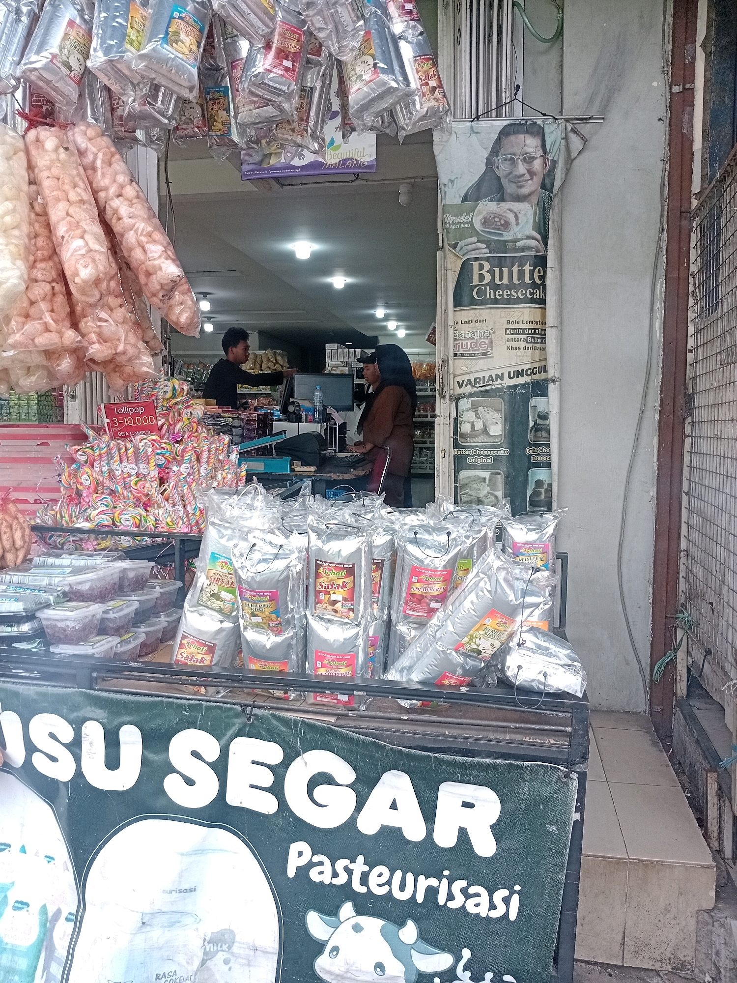 The image depicts the exterior of a shop in Lawang, Indonesia, likely a convenience store or small market, with a focus on snacks and beverages. In the foreground, a large green sign with white lettering reads "ISU SEGAR Pasteurisasi," indicating fresh pasteurized milk. The sign also features a stylized drawing of a cow. Behind the sign, a display rack is filled with numerous packaged lollipops of various colors and flavors. Above the lollipops, bags of snacks, possibly chips or crackers, are hanging. Further inside the shop, visible through an open doorway, two individuals are seen. One person, wearing a black shirt, appears to be working behind a counter with a computer monitor. Another person, wearing a brown hijab and a brown jacket, is standing near the counter. A poster is displayed on the wall to the right of the doorway, featuring a smiling person holding a slice of cake, with text below promoting "Butter Cheesecake" and "Banana Crudelt." The shop appears to be located on a street with a tiled walkway and a metal fence. The overall impression is a small, locally owned establishment selling a variety of goods.