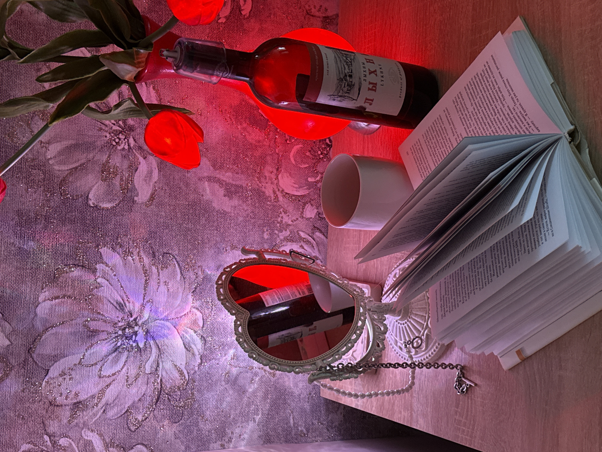 An overhead, slightly angled shot captures an intimate still life scene illuminated by a warm red glow. Artificial red tulips with green stems are arranged at the top left, alongside a dark glass bottle containing red liquid, likely wine. The bottle's label features text in Cyrillic, reading "ЛЫХНЫ" (LYKHNY), "Красное полусладкое" (Red semi-sweet), "вино натуральное" (natural wine), and partially "Product of abkhazia". This bottle rests on a red, circular object, which appears to be a light source casting the dominant red hue.
To the right, an open book lays flat, with its pages fanned out. Visible text in Russian includes character names like "Рейчел," "Коннор," "Саманта," and "Хоуп," along with words such as "мафия," "Вашингтон," and "школьный автобус." The page number "163" is also visible. A white mug is positioned near the book.
In the bottom left, an ornate, vintage-style mirror with a detailed frame reflects the red light, the wine bottle, and parts of the background. A pearl necklace with a silver chain and charm is draped nearby. The entire arrangement sits on a light wooden surface. The background consists of a textured fabric or wallpaper featuring a large, intricate floral pattern, subtly shimmering with glitter and illuminated by a mix of red and soft purple light, creating a dreamy and cozy atmosphere, suggesting an evening or nighttime setting.