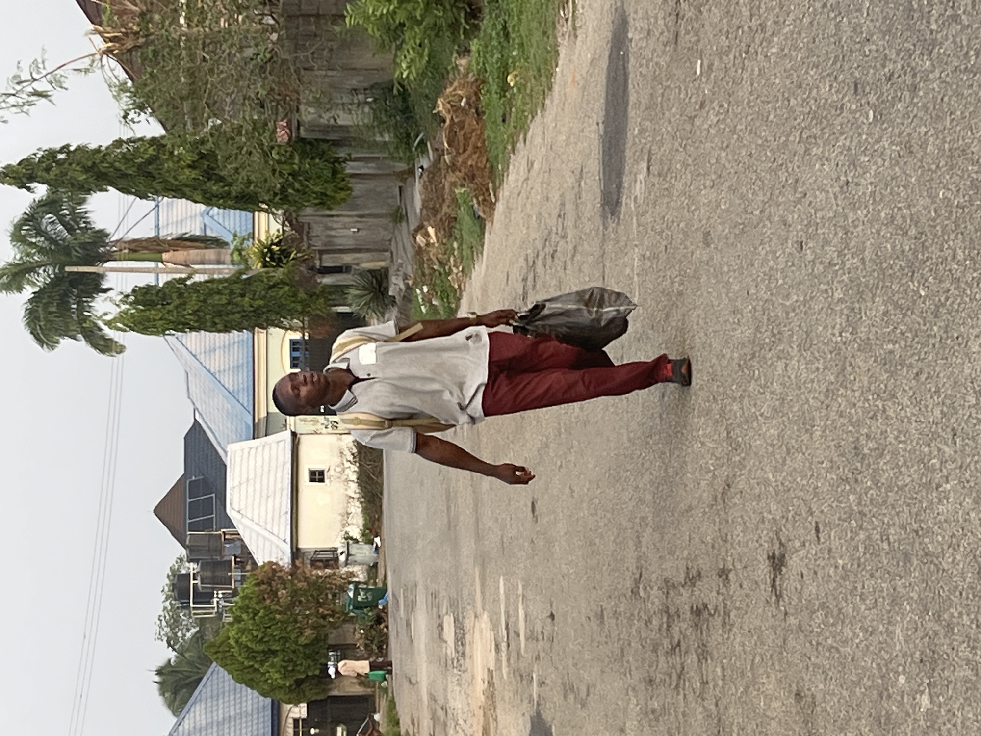 A man walks on a paved road in an urban environment. He wears a light-colored, short-sleeved shirt, dark red pants, and dark shoes. He carries a dark, possibly filled bag in his left hand. The road shows signs of wear and discoloration. To the side of the road is vegetation, including trees and shrubbery. In the background are buildings, some with dark roofs and water tanks, indicating a residential or mixed-use area. The sky appears overcast. The buildings suggest a planned residential area, common in Abuja, Nigeria. No specific landmark is discernible to pinpoint the exact location.
