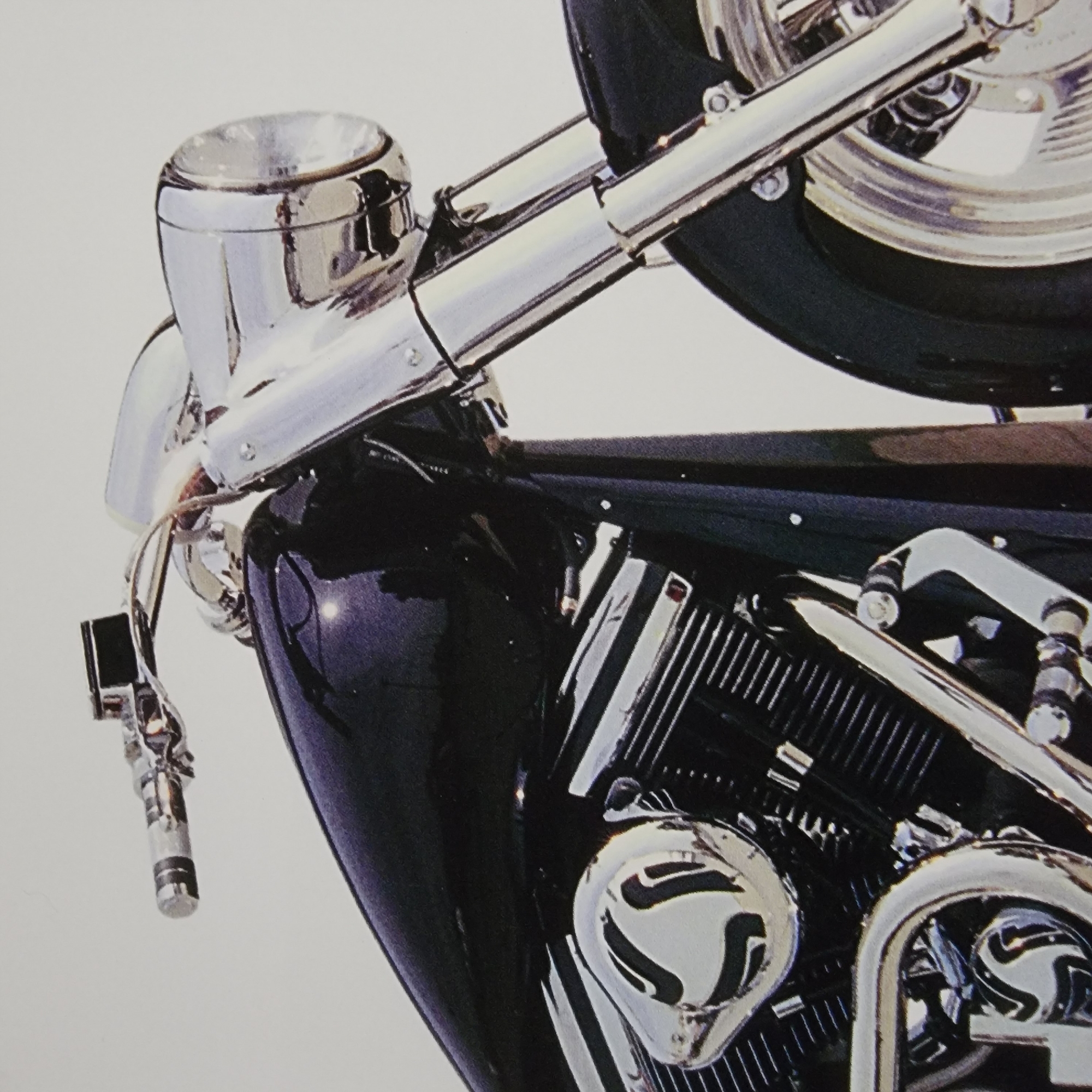 The image is a close-up, eye-level, medium shot of the front portion of a black motorcycle against a white background. The motorcycle features extensive chrome detailing on the handlebars, headlight, front forks, engine components, and exhaust pipes. A portion of the fuel tank, the front cylinder of the V-twin engine with a chrome cover, and the front tire are visible. The lighting is bright, creating reflections on the polished surfaces. There are no people or discernible environmental features related to a city.