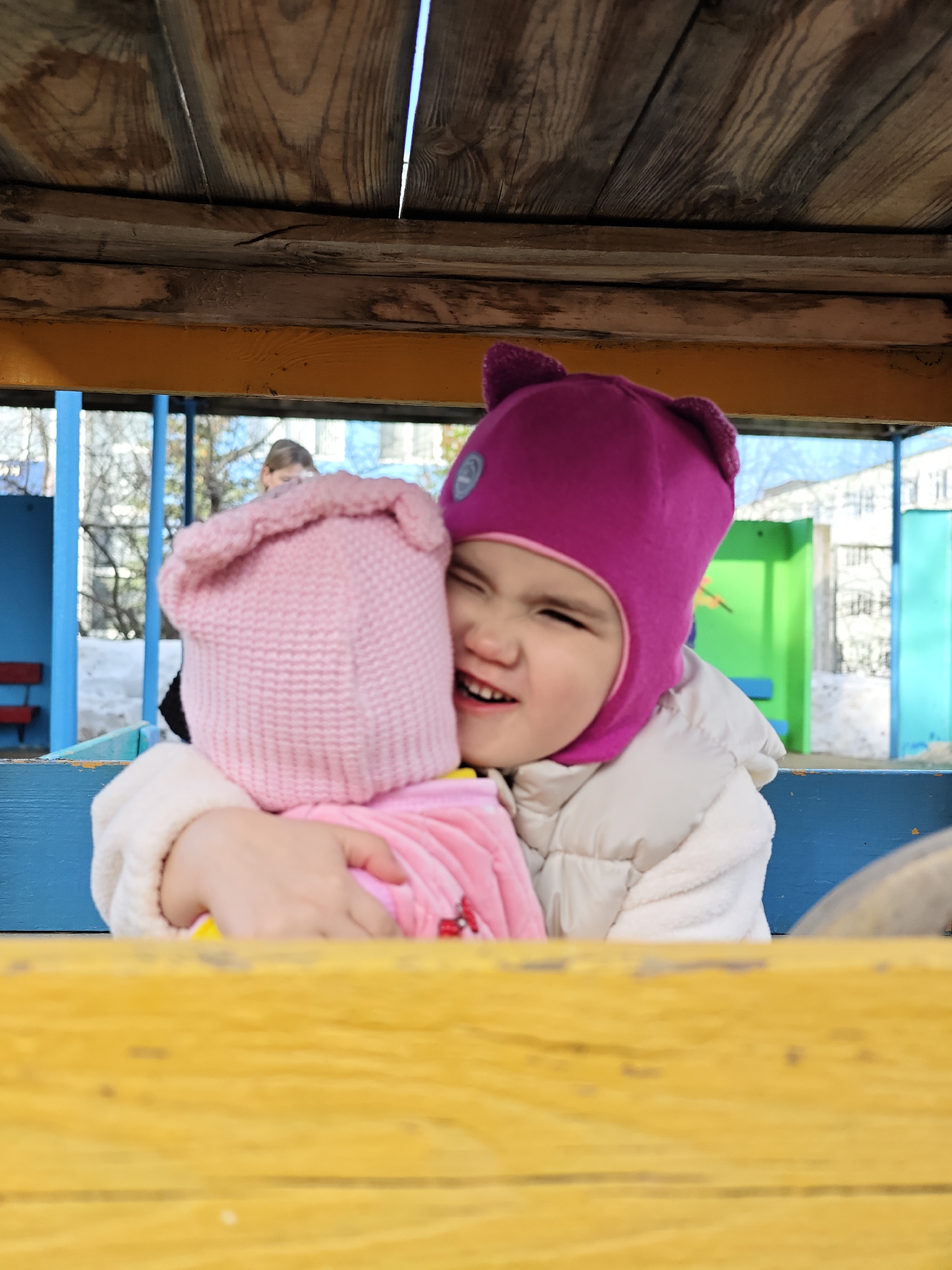A young girl, wearing a pink hat with ears and a white puffer jacket, is hugging a pink doll. The girl appears to be smiling or grimacing. The setting appears to be a children's play structure, with blue and yellow painted wood visible. The background shows a snowy outdoor environment. Based on prior knowledge, it is likely this image was taken in Chaikovsky, Russia.