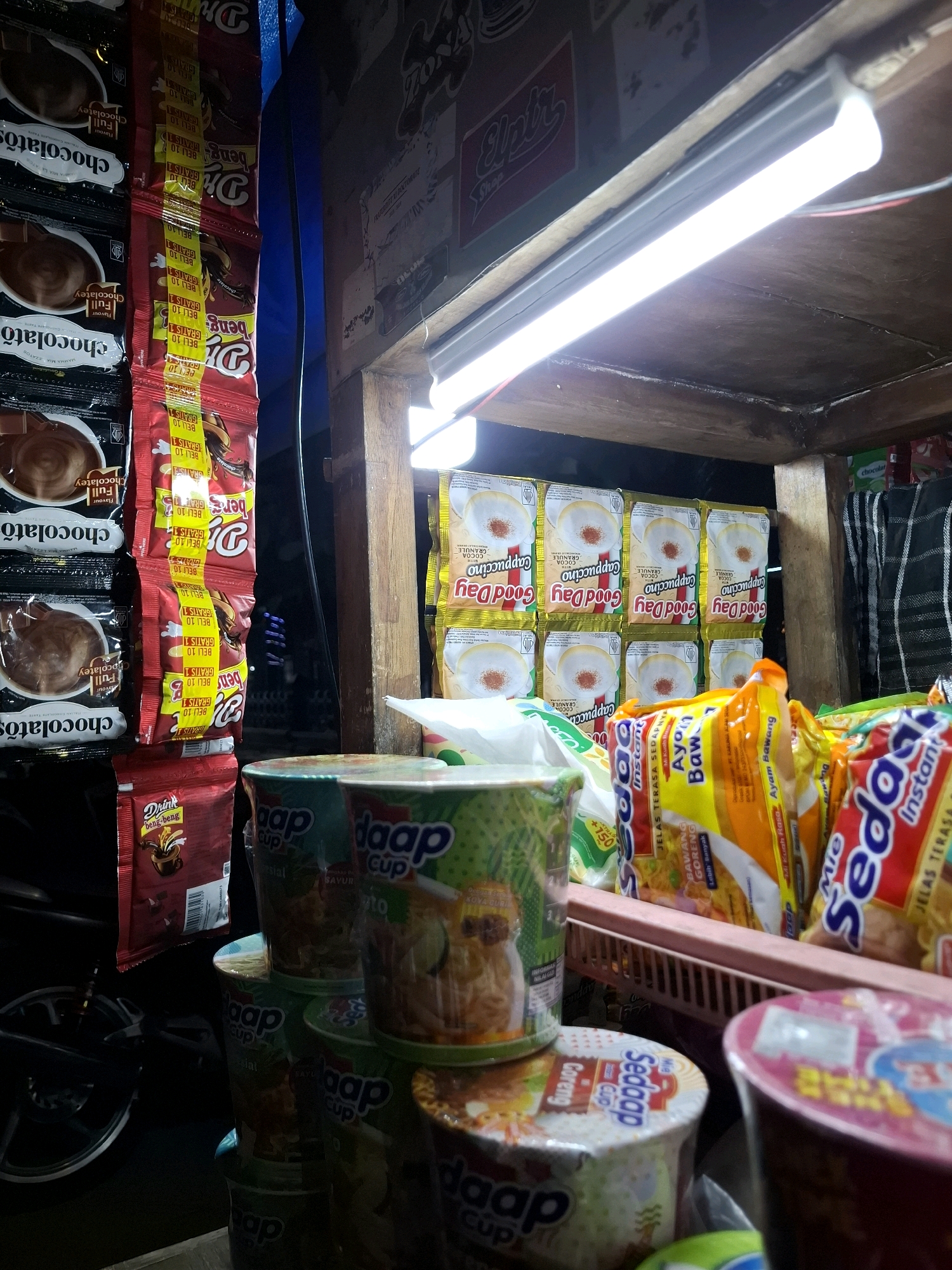 The image depicts a close-up, eye-level view of a street food stall, likely in the Pandean area of Indonesia, displaying a variety of packaged food items and instant noodle cups. The stall is illuminated by a bright fluorescent light fixture overhead.
Dominating the left side of the frame are several stacks of chocolate-flavored drink packets, some labeled "Full Chocolatey" and others with "Chocolatos." Adjacent to these, a red package of "Dring beng-beng" is visible.
To the right of the chocolate drink packets, rows of "Good Day Cappuccino" instant coffee sachets are neatly arranged, featuring images of a cappuccino. Further to the right, various instant noodle cups are stacked, including brands like "Daap Cup" and "Sedap Cup" with flavors such as "Mie Goreng Ayam Bawang" (fried noodles, chicken onion) and "Koya." The noodle cups are in different colors and designs, with some showing images of the prepared noodles.
A woven basket is positioned in the foreground, holding some of the instant noodle packages. The background shows the wooden structure of the stall and some black and white striped fabric. The overall scene suggests a retail environment for convenience food items at a street-side stall. No people are visible in the frame.