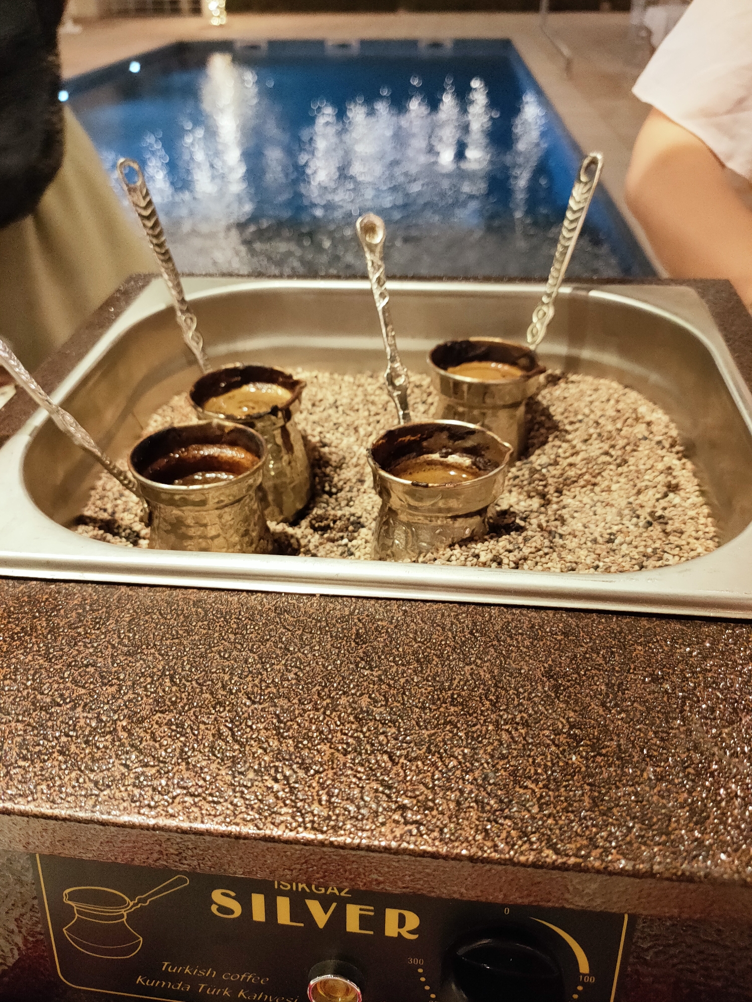 The image depicts a close-up view of a sand-filled apparatus used for brewing Turkish coffee. Four copper cezves (pots) containing coffee are submerged in the hot sand, with decorative spoons resting on the rim of the apparatus. The apparatus itself is a bronzed metallic unit with the brand name "ISIKGAZ SILVER" printed on its front panel, along with the text "Turkish coffee" and "Kumda Türk Kahvesi." A dial with numerical markings ranging from 0 to 300 is visible on the right side of the panel, suggesting temperature control. In the blurred background, a blue swimming pool with rippling water and some lights can be observed. The scene appears to be set outdoors, possibly on a terrace or poolside. No specific location in Cebalat, Tunisia, can be identified from the image itself, as it focuses on the coffee-making process rather than its geographical surroundings.