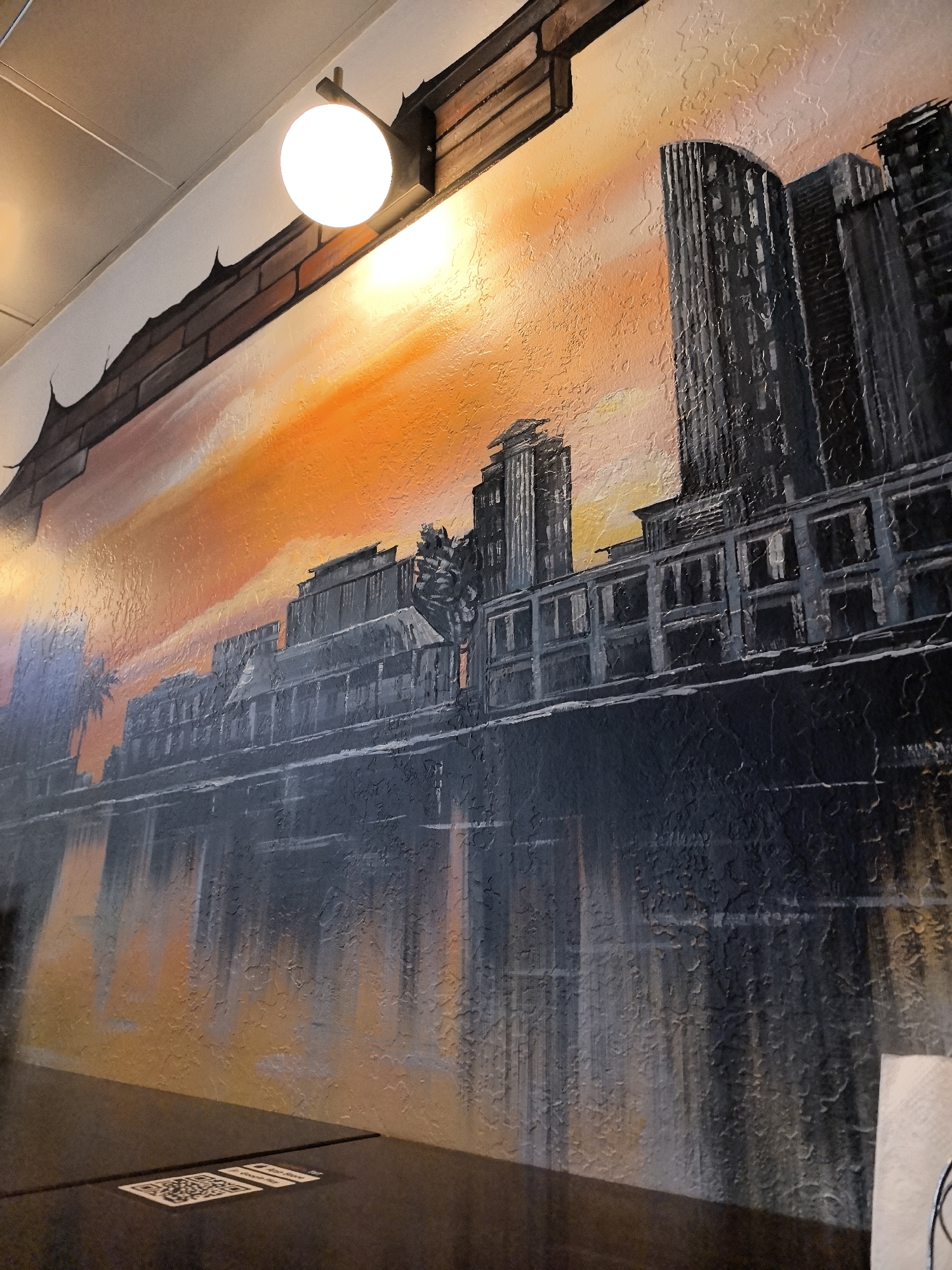 This is a photograph of a mural painted on a wall, depicting a city skyline at sunset. The mural features a row of dark, silhouetted buildings with various architectural styles, including modern skyscrapers and older, multi-story structures. The sky is a vibrant orange and yellow gradient, suggesting the setting sun. A body of water, likely a river or harbor, is depicted in the foreground, reflecting the colors of the sky and the buildings. The mural has a textured finish, giving it a tactile appearance. Above the mural, a portion of a brick wall is visible, with a light fixture attached to it. The light fixture is a simple globe light with a metallic bracket. The overall scene is an artistic representation of an urban landscape.