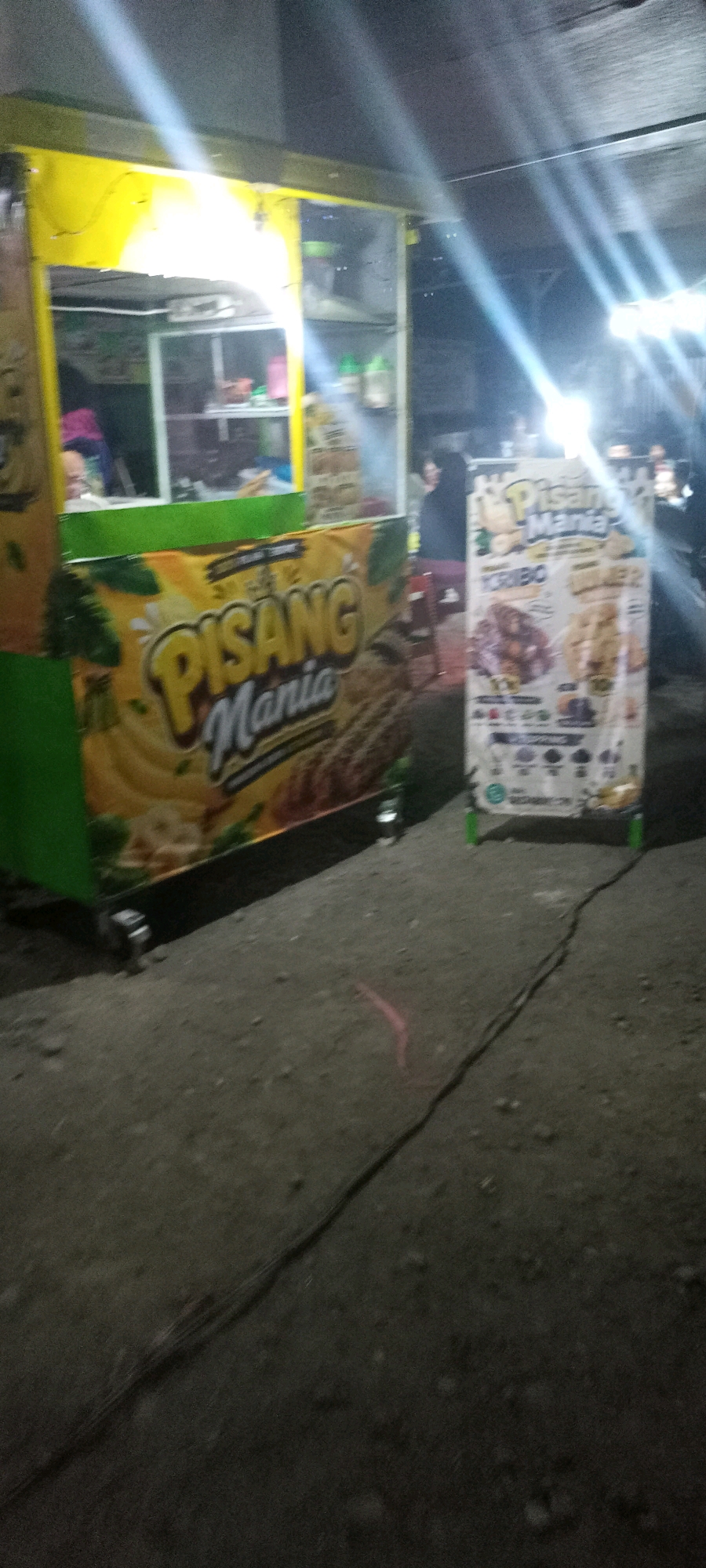 The image shows a street food cart at night in Bendotretek, Indonesia. The cart is primarily yellow and green and displays signage for "Pisang Mania." One sign features images of fried bananas. A second, taller sign to the right also advertises "Pisang Mania" and lists different menu items, including "YORIBO" and "JUICE 2." The cart appears to be illuminated by bright overhead lights, creating strong reflections and glare. The ground in front of the cart is a dark, rough surface, possibly dirt or gravel, with a black cable running across it. In the background, blurred figures and the outline of other stalls or buildings are visible. The overall scene suggests a busy night market or street food area.