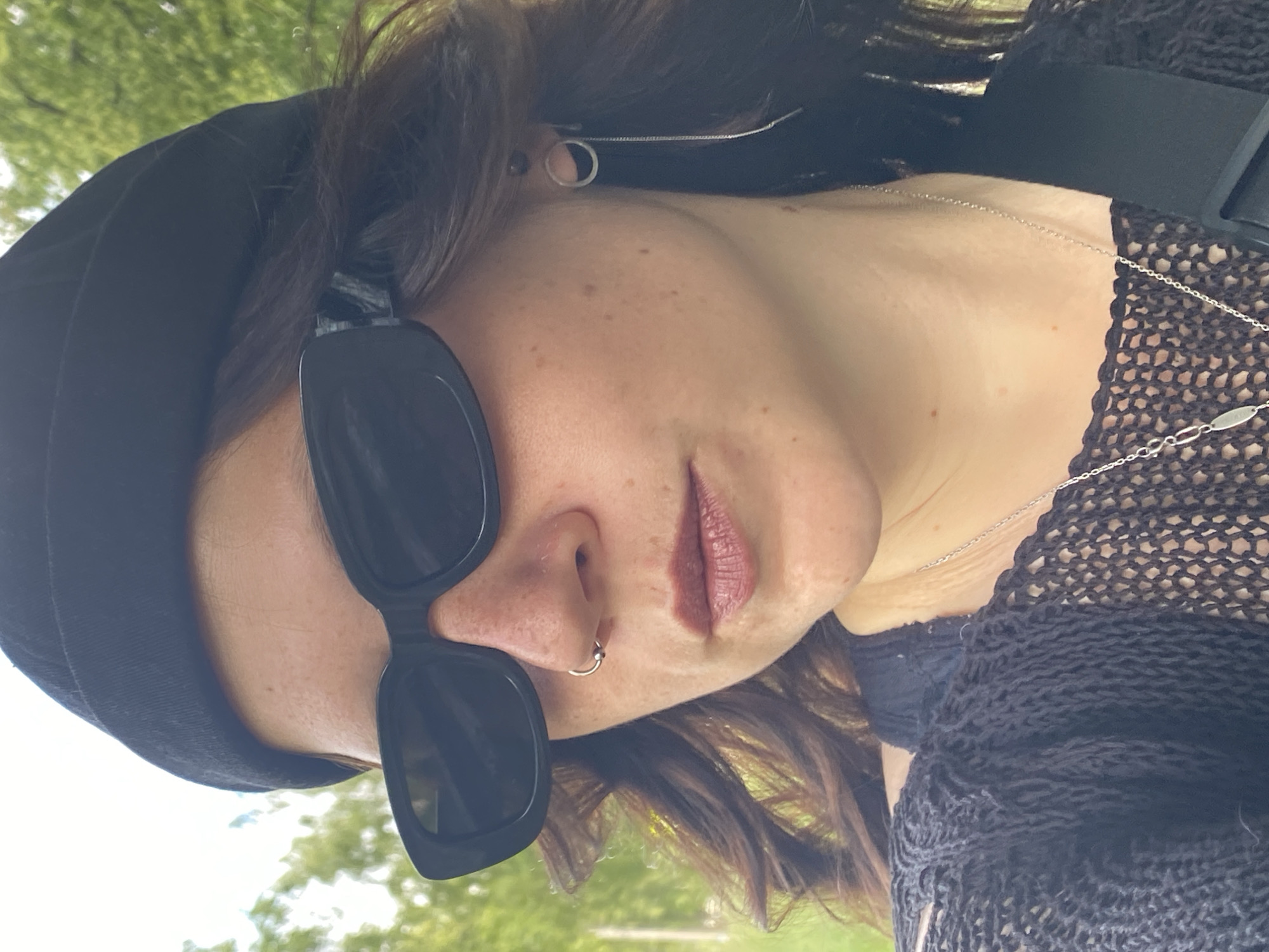 A medium shot captures a woman wearing a black beanie, black sunglasses, and a black mesh cardigan. She has a nose ring and a long chain earring. The background consists of blurred green foliage, suggesting an outdoor setting, possibly in Chaikovsky, Russia, given its natural landscapes.