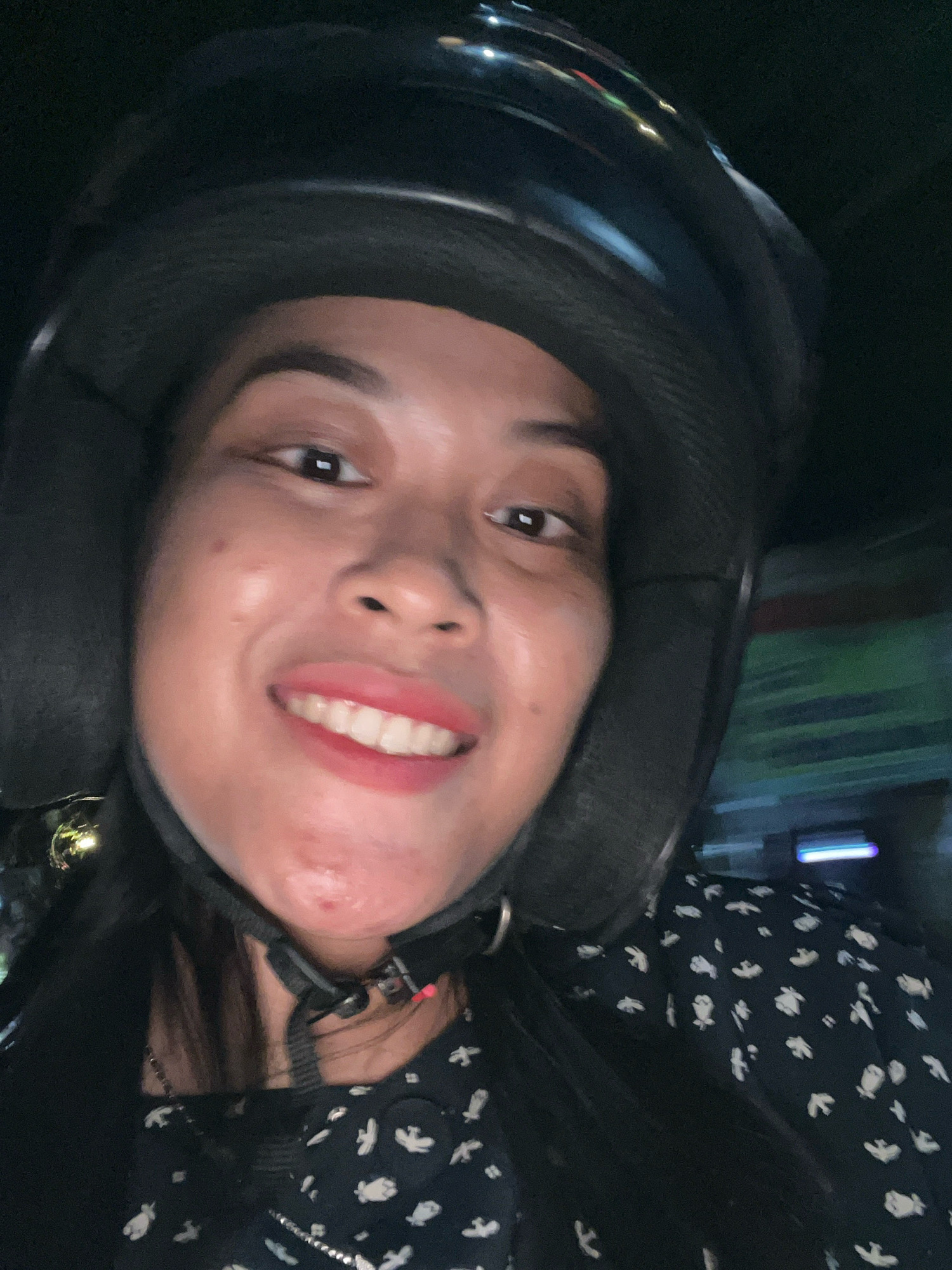 A young woman wearing a black helmet and a black patterned dress is shown from the chest up, taking a selfie at night in what appears to be a moving vehicle in the city of Kecamatan Pati, Indonesia. The woman has dark hair, brown eyes, and is smiling, revealing her teeth. She is wearing pink lipstick. The background is blurred, suggesting motion. The lighting is dim, with some reflections on the helmet.