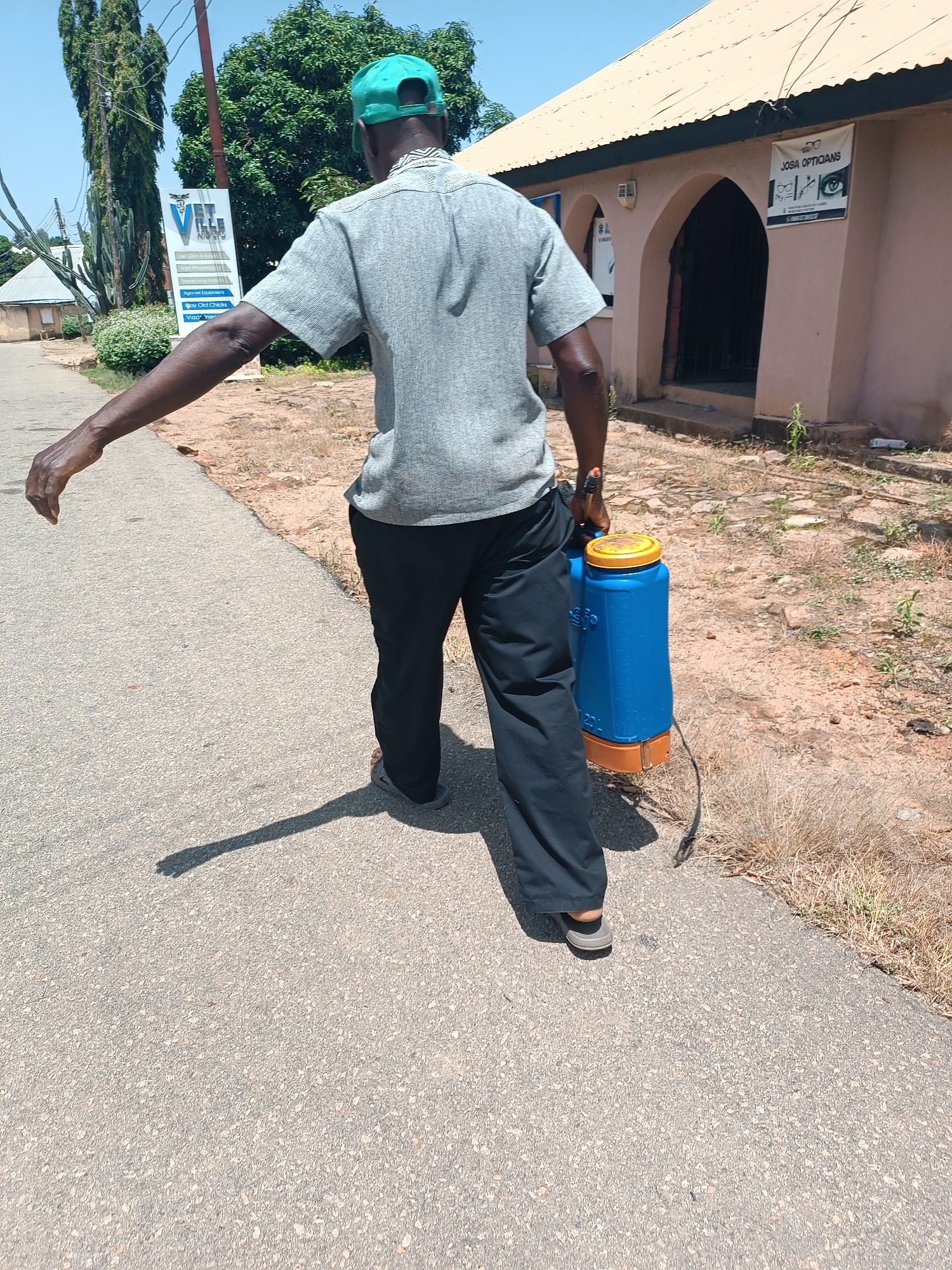 In Jos, Nigeria, a person wearing a grey short-sleeved shirt, black trousers, and a green cap walks along an asphalt road, carrying a blue and orange sprayer. The person's right arm is extended to the side, and their shadow is cast on the road. To the left of the road, a white sign with "VELL CLINIC & SUPPLIES" and other text is visible next to a tall, thin tree and some dry grass. Behind the person, a light-colored building with an arched entrance and a light brown roof is situated. A banner with "JOSA OPTICIANS" and an eye graphic is attached to the building. The environment appears dry and sunny, with sparse vegetation and loose soil.