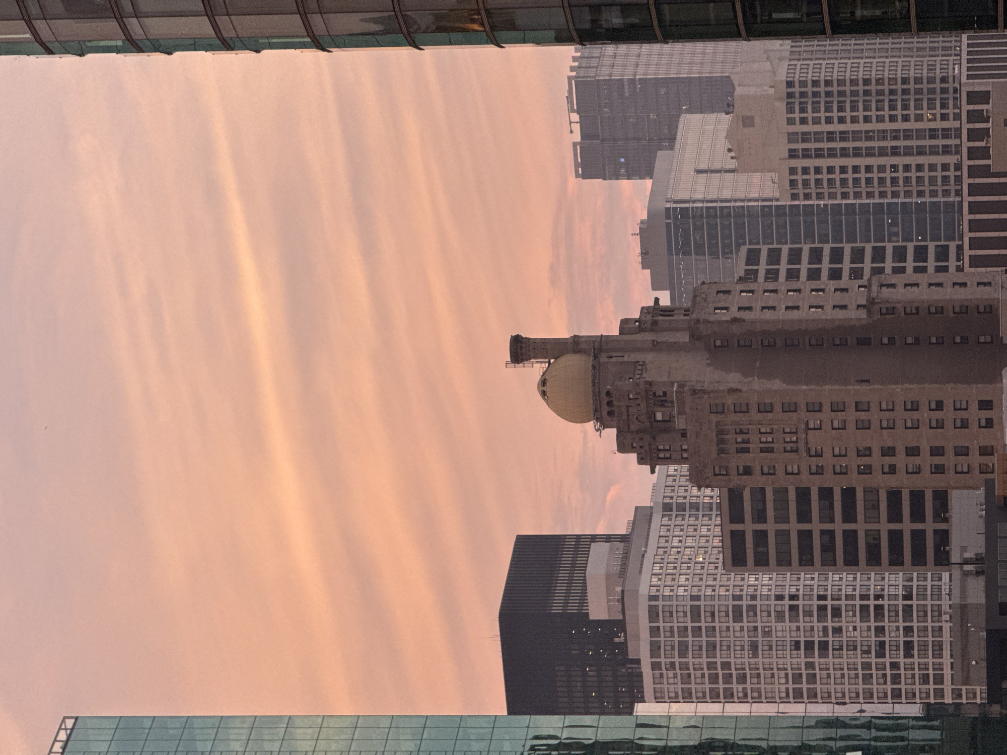 The image shows a cityscape with multiple buildings under a pale pink sky with wispy clouds. In the foreground, a tall, light brown building with a distinctive golden dome on top is prominent. To its left, a modern, dark-colored building with many windows stands out. Further to the left, a glass-covered skyscraper with a greenish tint is visible. On the right side of the frame, another modern glass-covered skyscraper curves. In the background, more buildings of varying heights and architectural styles fill the view. The scene appears to be taken during either sunrise or sunset, given the soft lighting and the color of the sky. The image does not contain any people. The overall impression is of an urban environment during twilight.