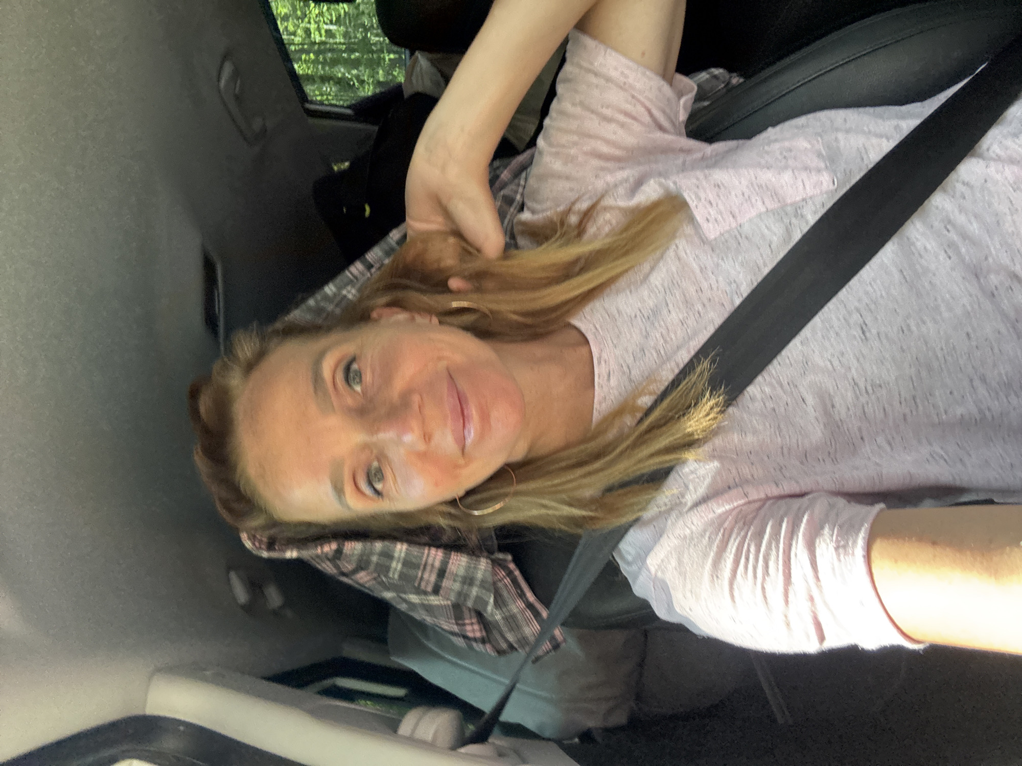 A woman with blonde hair, wearing a light pink t-shirt and a seatbelt, is in the driver's seat of a car. She is smiling and touching her hair. The car is packed with luggage and blankets, suggesting a road trip or moving. Outside the car window, trees are visible, indicating a rural or forested area. The image was taken in Portovoe, Russia.