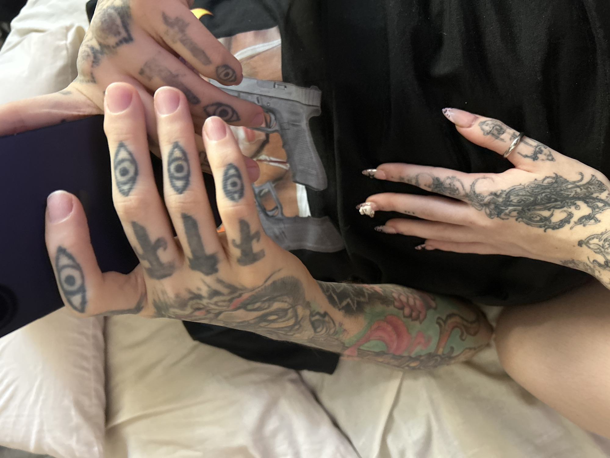 A close-up, high-angle shot captures the hands and forearms of two individuals in an indoor, domestic setting, likely a bedroom, indicated by white/cream bedding and pillows.
The person on the left holds a dark purple or blue smartphone. Their hands and left forearm are extensively tattooed. Distinctive tattoos include multiple stylized "eye" designs on the fingers, dagger-like symbols on the back of the hand, and a large, colorful sleeve tattoo on the forearm featuring intricate motifs, including what appears to be an animal head (possibly a fox or wolf) integrated with vibrant pink and green organic patterns. Their fingernails are natural and unpolished.
The person on the right displays their right hand and forearm, which are also heavily tattooed. The back of this hand features an elaborate, flowing blackwork design. A simple silver band ring is worn on the ring finger. Their fingernails are long, almond-shaped, and adorned with a light polish and subtle decorative elements, possibly glitter or rhinestones. This individual is wearing a black short-sleeved top, which features a printed image of a realistic grey handgun, resembling a Glock pistol.
The hands are positioned in close proximity, suggesting a relaxed interaction or shared space. No text is visible within the image. While the location context is Buenos Aires, Argentina, no specific visual cues within the image confirm this.
