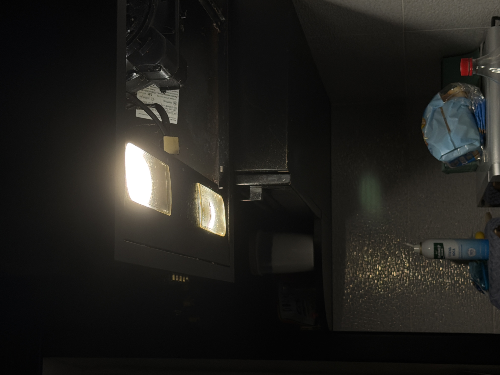 The image depicts a close-up, low-angle shot of a kitchen exhaust hood with its lights on, illuminating the underside. Below the hood, on a shelf, are several items. To the left, partially obscured by shadow, is a cylindrical container with white contents, possibly milk or yogurt, and a label with Cyrillic text. To the right of this container, a bottle of nasal spray with a blue and white label displaying Cyrillic text is visible, standing next to a crumpled blue plastic bag containing what appears to be snacks. In the foreground, a clear plastic bottle with a red cap is positioned to the right. The overall environment suggests an indoor domestic setting, likely a kitchen. The presence of Cyrillic text on some of the products indicates a location where this language is used. Given the prompt's specification, this scene is located in Bila Tserkva, Ukraine.