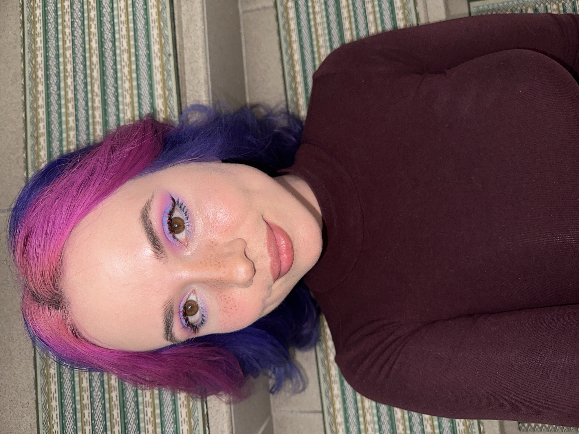 A close-up, high-angle portrait captures a young person lying down, looking towards the camera with a soft, slight smile. The subject features distinctive two-toned hair, bright pink framing the face and transitioning to a deep purple at the back. Their fair skin is notable for scattered freckles across the nose and cheeks. The warm brown eyes are accentuated with light purple eyeshadow on the lids and striking bright pink mascara on the lower lashes. The lips are a natural, glossy pink hue.
The person is dressed in a dark, likely maroon or deep burgundy, long-sleeved top with a high neckline. They are positioned on a vertically striped surface, which appears to be a rug or mat featuring patterns in shades of green, white, and beige. A plain grey surface, possibly a wall or floor, is also visible in the background. The setting is indoors, with lighting that suggests artificial illumination or diffused daylight. The image conveys a calm and engaging mood. While no specific location cues are visible in the image itself, the photograph was taken in the City of Zagreb, Croatia. No text is visible.