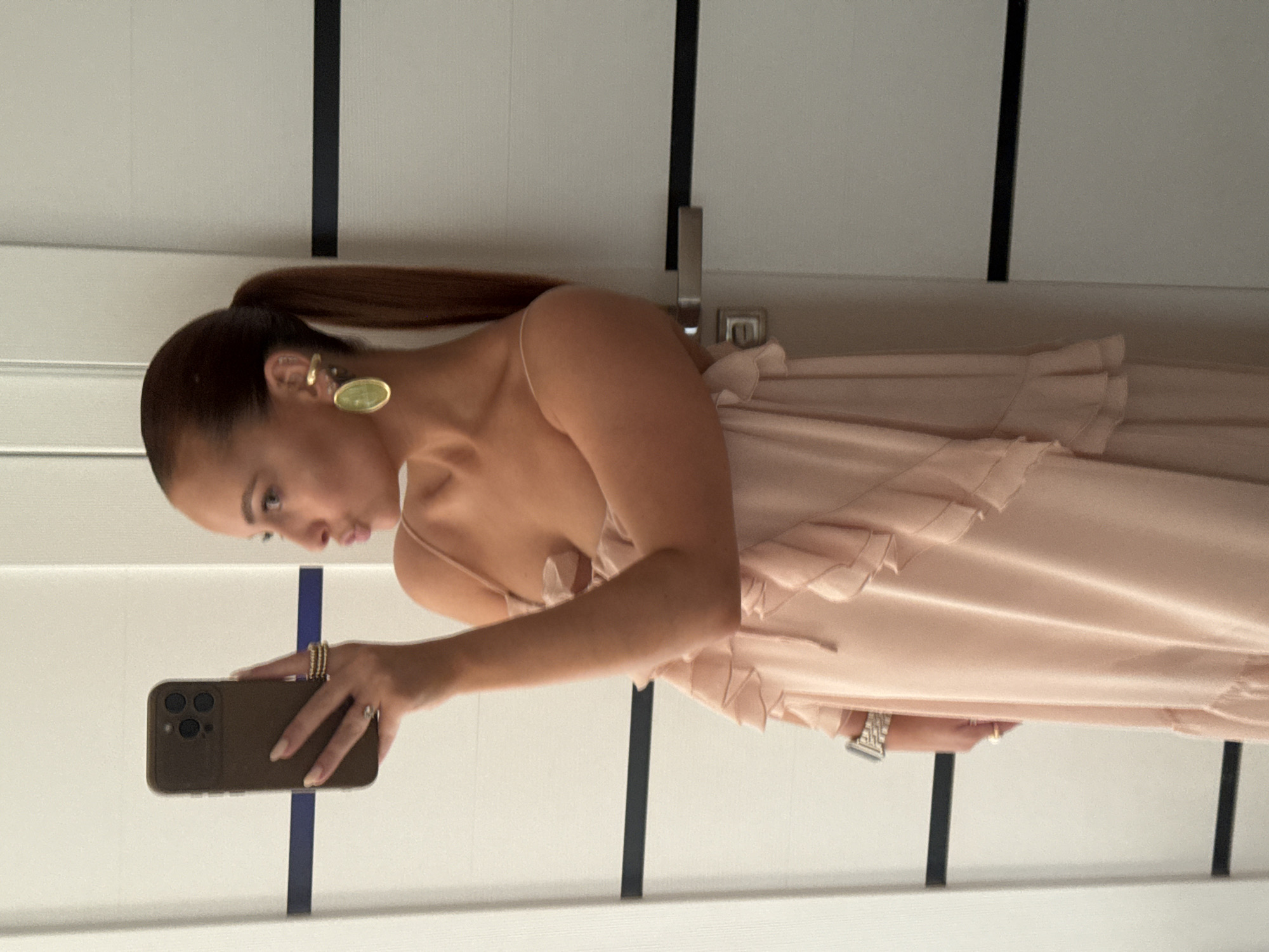 A woman is taking a selfie in front of a door or a wall with vertical dark lines. She has brown hair pulled back in a ponytail and is wearing a light pink dress with ruffles, as well as gold jewelry. The phone she's using is an iPhone with a brown case. The location is possibly indoors but cannot be confirmed as Chaikovsky, Russia based on visual details.