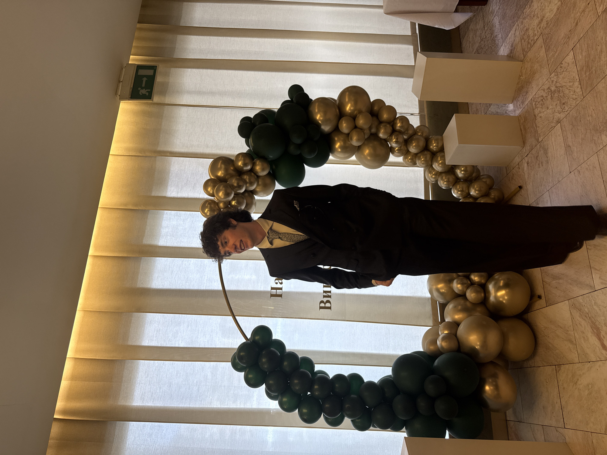 The image shows a person in a dark suit standing in front of a circular balloon arch. The balloon arch is decorated with gold and dark green balloons and is in front of a window with blinds. The person is smiling and looking off to the side. There are also white rectangular podiums nearby. The background appears to be an indoor event space. The balloon arch has the phrase "На Ви" written on it. Based on this phrase, it is highly likely that this photo was taken in Bulgaria, as that's the official language. Without further information, pinpointing the specific location in Sofia is not possible.