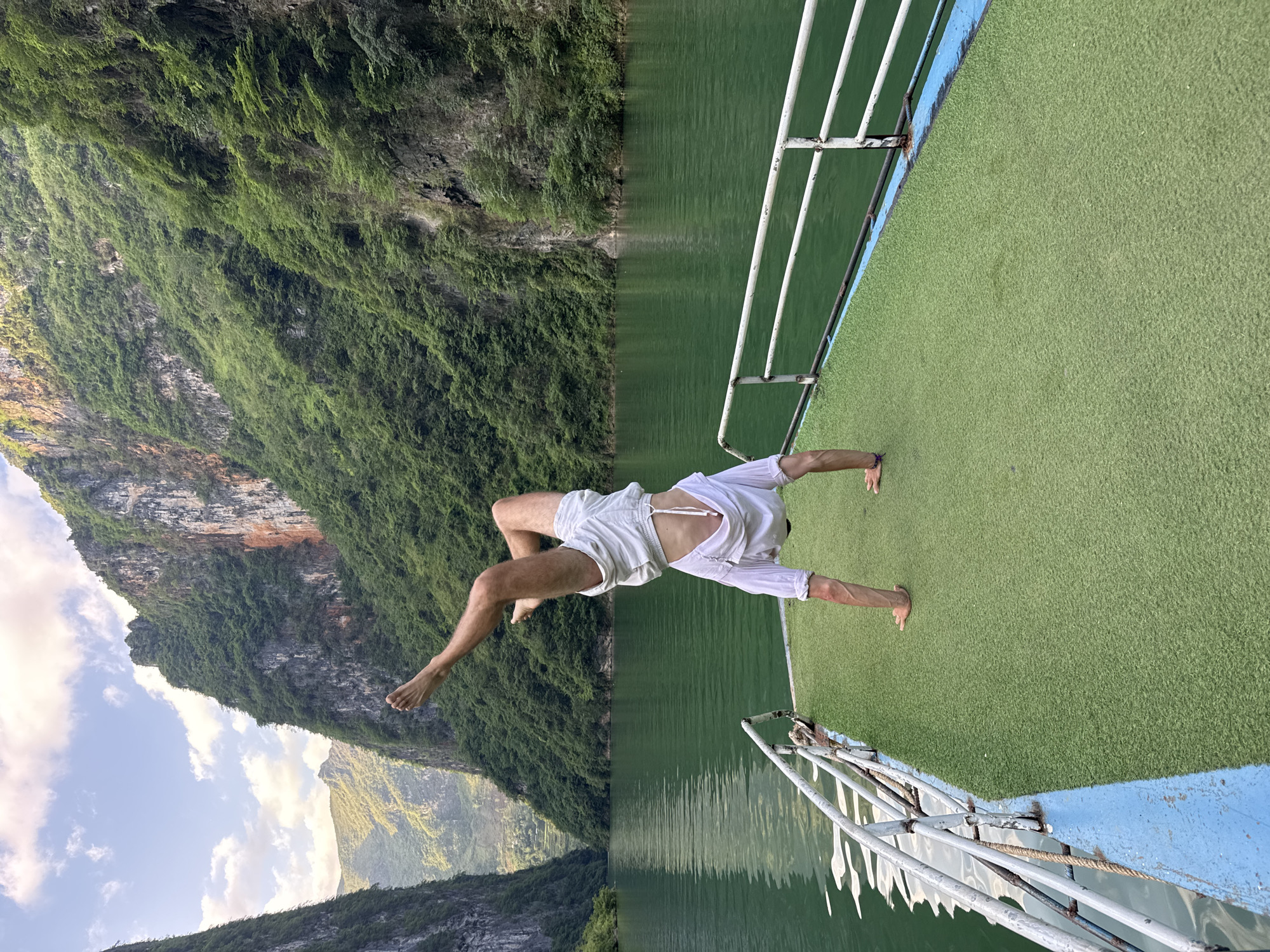 A man is performing a handstand on the edge of a boat. The boat is located on a body of water surrounded by cliffs covered in green vegetation. The location is the city of Đồng Văn Commune, Vietnam. The man is wearing white shorts and no shirt. The boat has a green artificial turf floor and white railings. The sky is visible in the background, with some clouds.