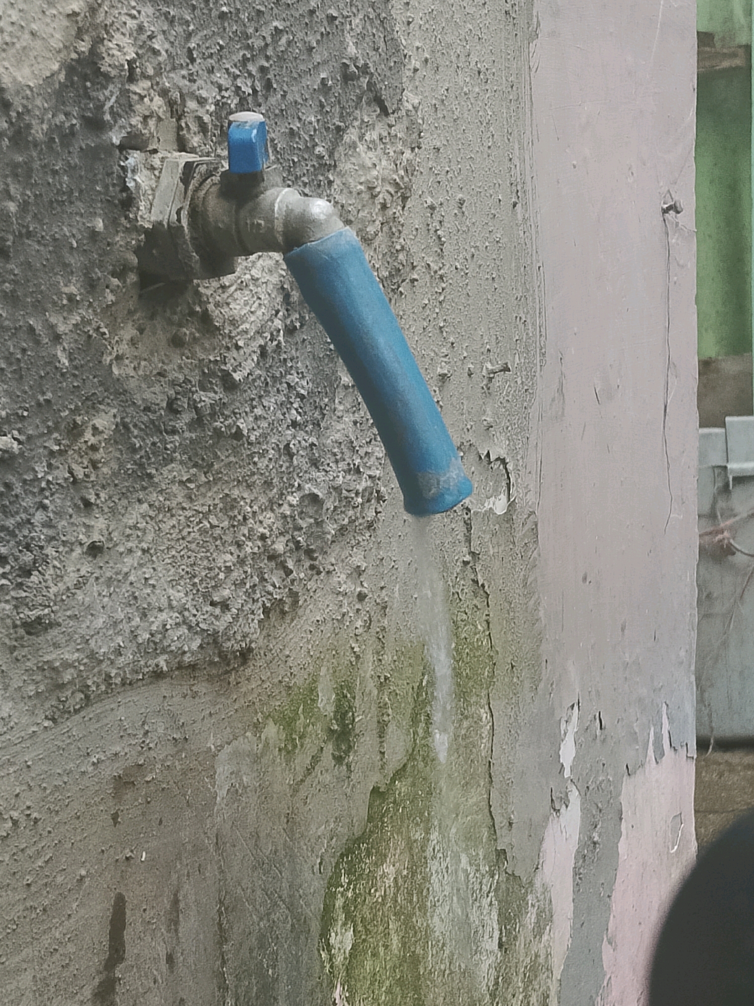 A close-up, outdoor, eye-level shot captures a water faucet mounted on a textured, grey concrete wall. The faucet is made of metal, with a blue handle and a blue-wrapped spout from which a thin stream of water is flowing downwards. The wall itself is rough and uneven, showing signs of wear and patches of green moss or algae. To the right of the faucet, the wall transitions to a smoother, light-colored surface, also with peeling paint. In the background, a glimpse of a green vertical structure and a white metal object can be seen. The image is shot from a static perspective, with the water stream appearing slightly blurred due to motion.