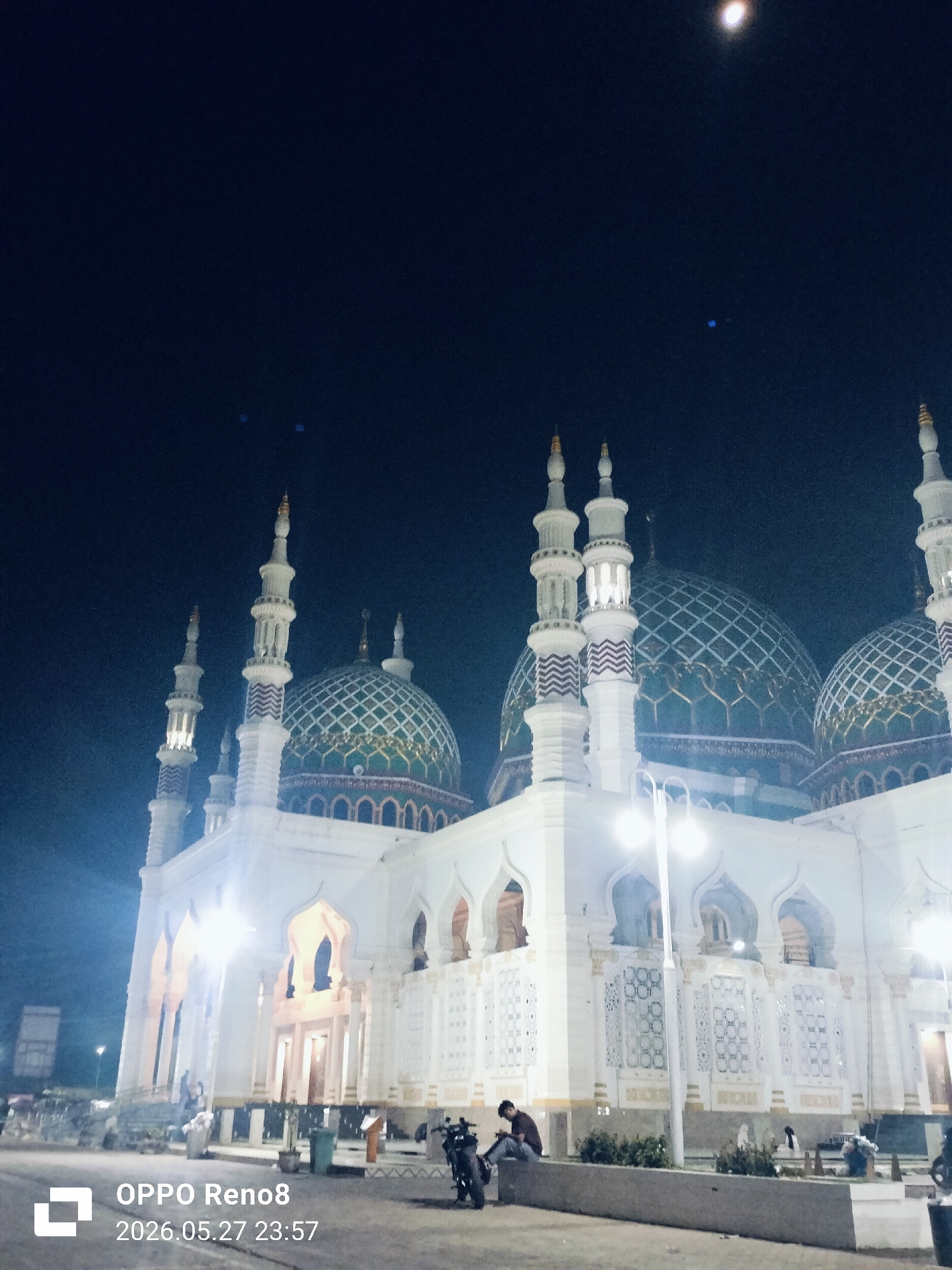 The image depicts the Baiturrahman Grand Mosque at night in Kotapantonlabu, Indonesia. The mosque features white walls, arched windows, and multiple domes decorated with a green and gold lattice pattern. Several minarets with white and grey striped patterns rise from the structure. Illumination from street lights and the mosque's internal lighting casts a bright glow on the building's facade. In the foreground, a street scene includes a motorbike and a person sitting on a low wall in front of the mosque. The night sky is dark, with a visible moon. The image includes a watermark indicating "OPPO Reno8" and the date "2026.05.27 23:57."
