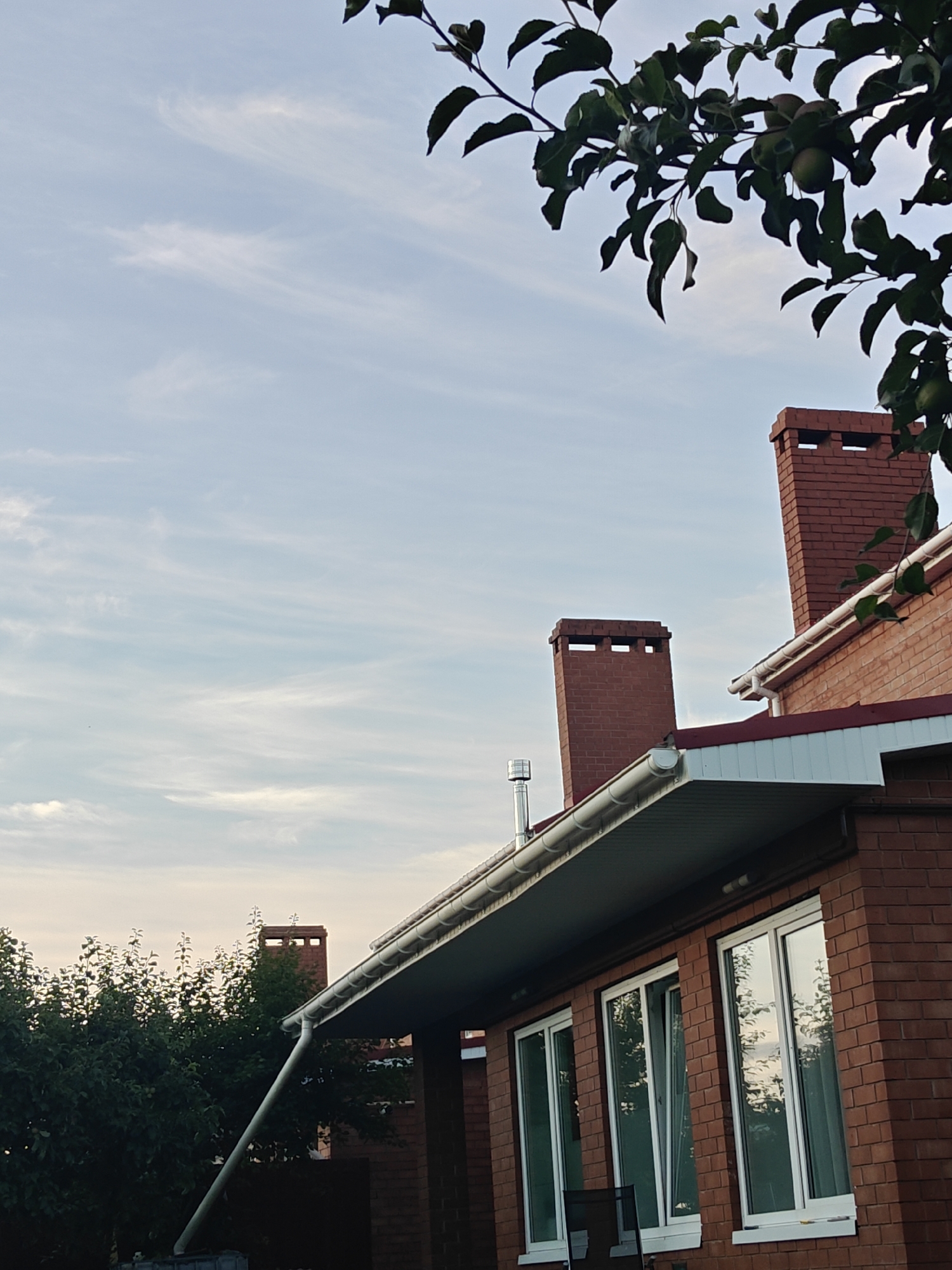 The image captures an exterior view of a red brick house in Podstyopki, Russia, under a partly cloudy sky. The house features white-framed windows and a white rain gutter system. Two brick chimneys are visible on the roof, one taller than the other. A tree branch with green leaves and what appear to be small fruits hangs in the upper right portion of the frame, partially obscuring the sky and the roof. The background shows dense green foliage from trees. The lighting suggests it is either early morning or late afternoon.