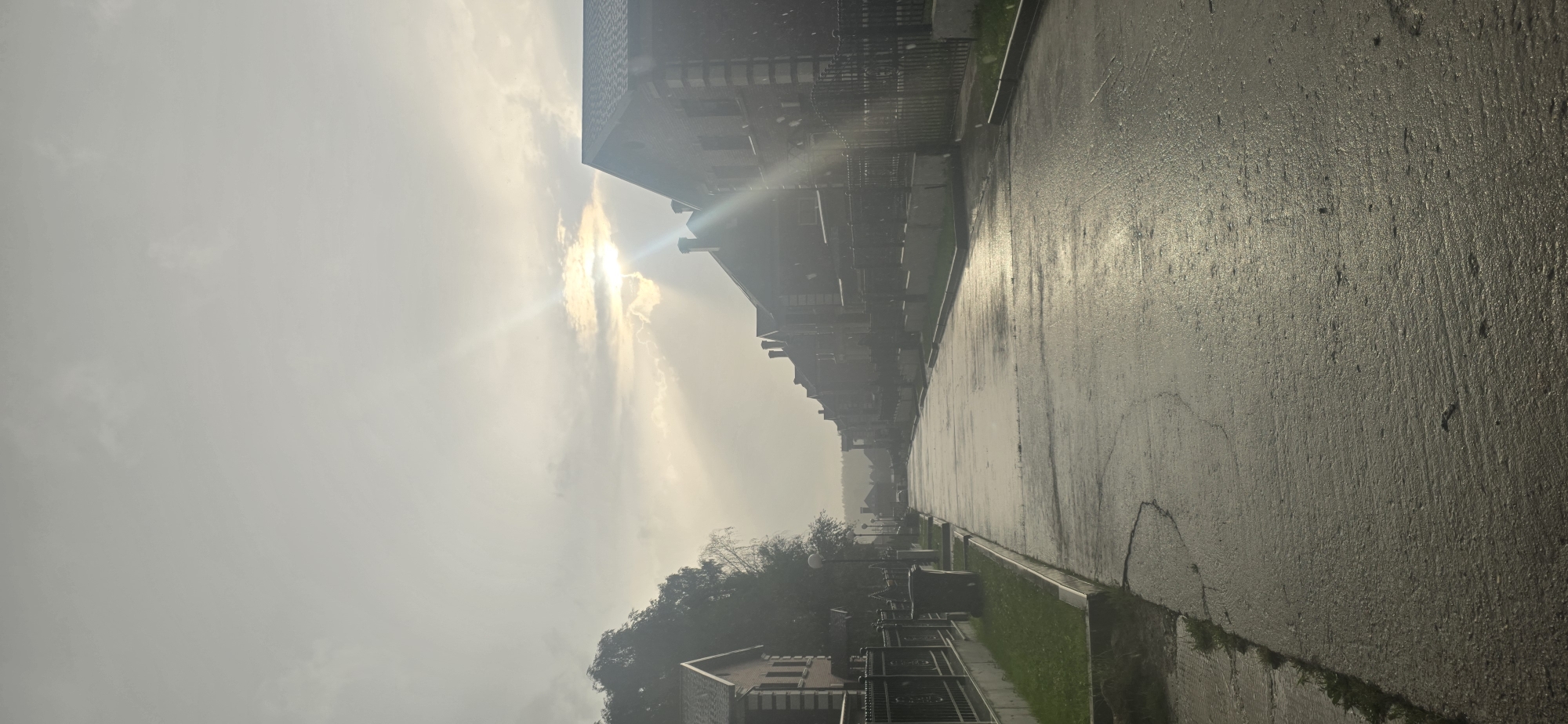 A vertical shot captures a street in the village of Русская Деревня-2 Кп, Russia, under an overcast sky with sunbeams piercing through the clouds. The wet asphalt road, reflecting the dull light, stretches into the distance, lined with identical brick houses with dark roofs and ornate black fences. A line of trees is visible on the left side of the street. The scene appears to be captured during or shortly after a rainfall, with small white specks, possibly raindrops or snowflakes, visible in the air. A gray trash can sits on a patch of grass next to the sidewalk on the left. The overall atmosphere is subdued and possibly cool, with the muted tones of the sky and the reflective wet road contributing to a somber mood.