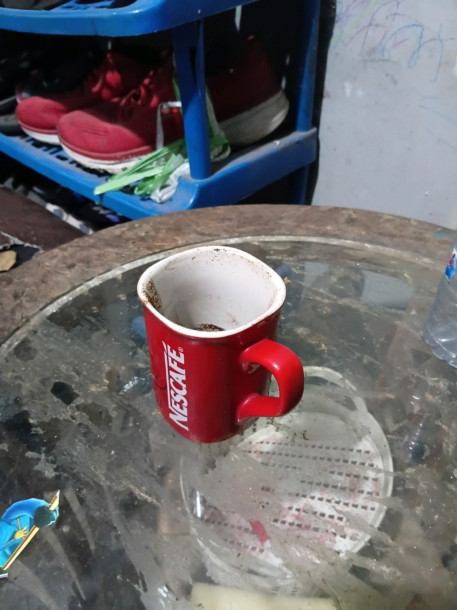 A red Nescafé mug containing a small amount of coffee grounds sits on a dirty glass table. In the background, a blue shoe rack holds red sneakers. The scene appears to be indoors, possibly in a residential area in Cikal, Indonesia, given the context of the provided information. The overall environment is cluttered and appears neglected.