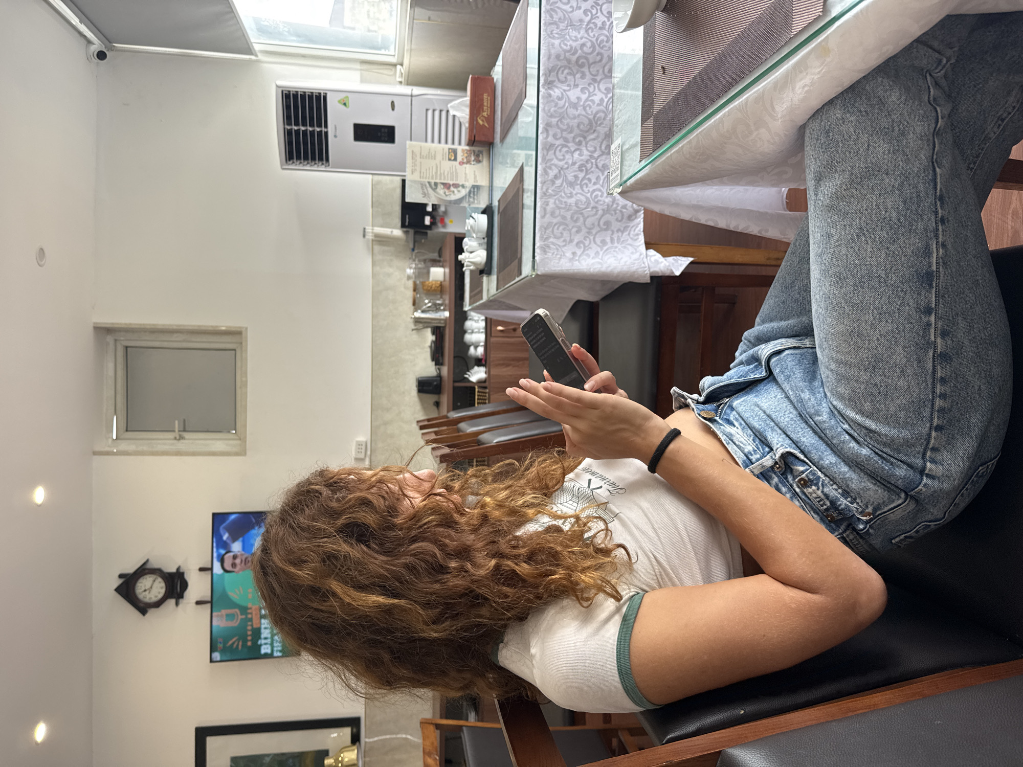 A young woman with long, curly brown hair is sitting at a table in a restaurant in Hanoi, Vietnam. She is wearing a white crop top with green trim and light blue jeans. She is holding a smartphone in her hands and looking at it. The restaurant appears to be casual with wooden chairs and tables covered with white tablecloths. There is a television screen in the background displaying an image of a man. An air conditioning unit is visible on the right side of the image. The overall scene is one of a relaxed dining environment.