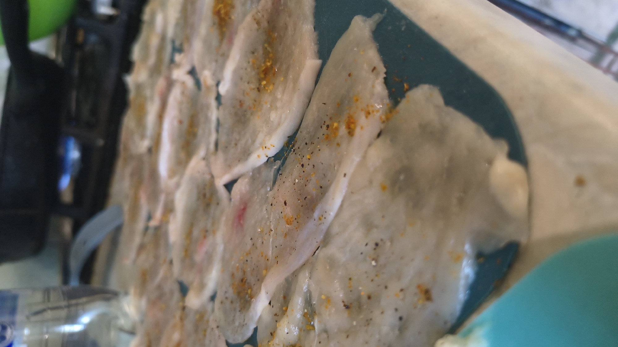 The media file displays a close-up, tilted view of several thin, translucent slices of food, possibly fish or thin cuts of meat, arranged on a teal-colored surface. The food items are sprinkled with what appears to be a mix of spices, including black pepper and a yellowish-orange spice, likely paprika or turmeric. Some pieces show slight pinkish hues, suggesting they might be raw or lightly cooked.
In the background, to the left, a dark, metallic object is visible, with a bright blue flame emanating from it. This could be a cooking apparatus, such as a stove burner or a torch. A green spherical object is also partially visible in the upper left corner. To the bottom left, a clear plastic bottle with some liquid inside and reflective elements is present.
The lighting suggests an indoor setting, potentially a kitchen or food preparation area. There are no discernible people in the frame. The overall impression is of food being prepared or presented for consumption. The specific location context provided is Vinnytsia, Ukraine, but no visual cues within the image directly support or contradict this. There is no visible text in the image. The focus is entirely on the food and the immediate preparation environment.