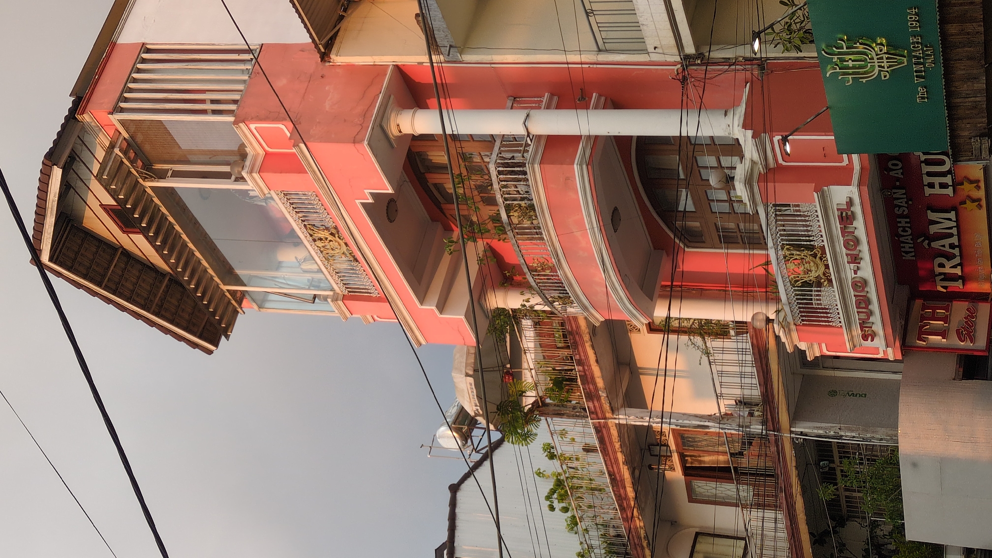 This photograph depicts a building with an elaborate facade, which is identified as "Studio Hotel" and the "Vintage 1994," located on Nguyễn Văn Trỗi street in Da Lat, Vietnam. The building has a prominent pink exterior with architectural details such as balconies, decorative railings, and a white supporting column. Signage in Vietnamese script is visible, advertising "KHÁCH SẠN - ÁO TRẦM HƯ" with a "TH Store" below. Power lines are entangled in the foreground, and the sky is overcast, indicating either dusk or dawn.