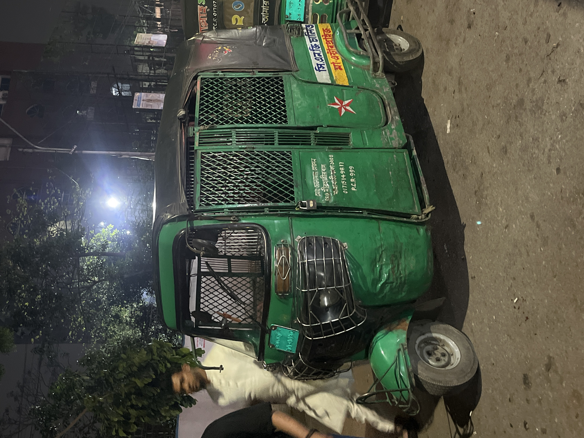 The image depicts a CNG auto-rickshaw in Dhaka, Bangladesh, illuminated by streetlights at night. The vehicle is green, with a caged passenger area. Text on the auto-rickshaw includes contact information and the phrase "সি. এন.জি চালিত," indicating it's CNG-powered. A partial view of people is visible at the bottom of the frame. The setting appears to be an urban street, with buildings visible in the background.