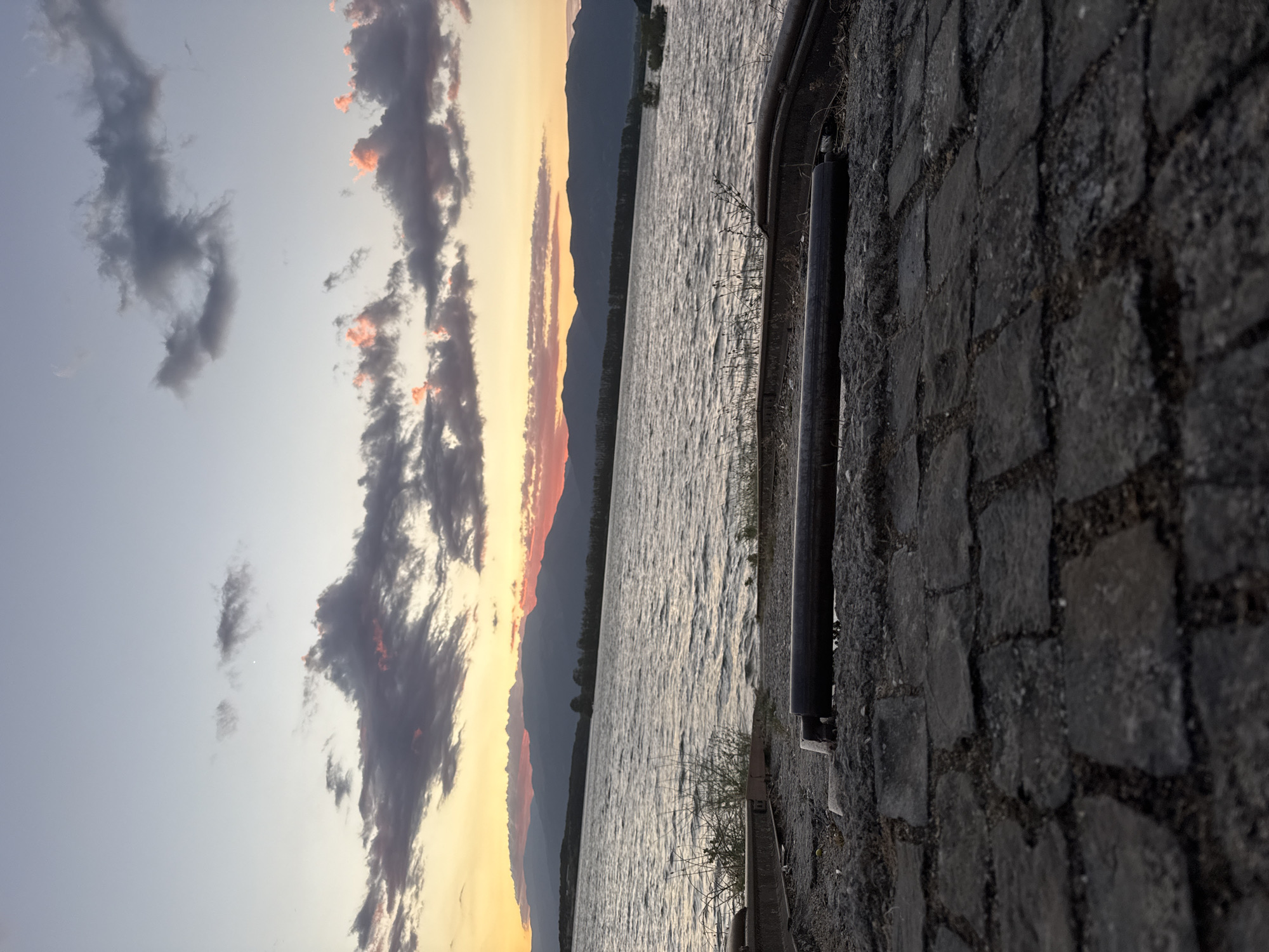 The image depicts a sunset over Koprinka Reservoir in Koprinka, Bulgaria. In the foreground, there are old cobblestones and what appear to be railroad tracks, suggesting a disused industrial area or pier. The reservoir itself is choppy with visible ripples and waves. Across the water, a distant treeline borders the foothills of mountains, silhouetted against the vibrant orange and pink hues of the setting sun. The sky is a gradient of light blue to a pale yellow, with scattered clouds, some tinged with pink from the sunset. There are no people visible in the image.