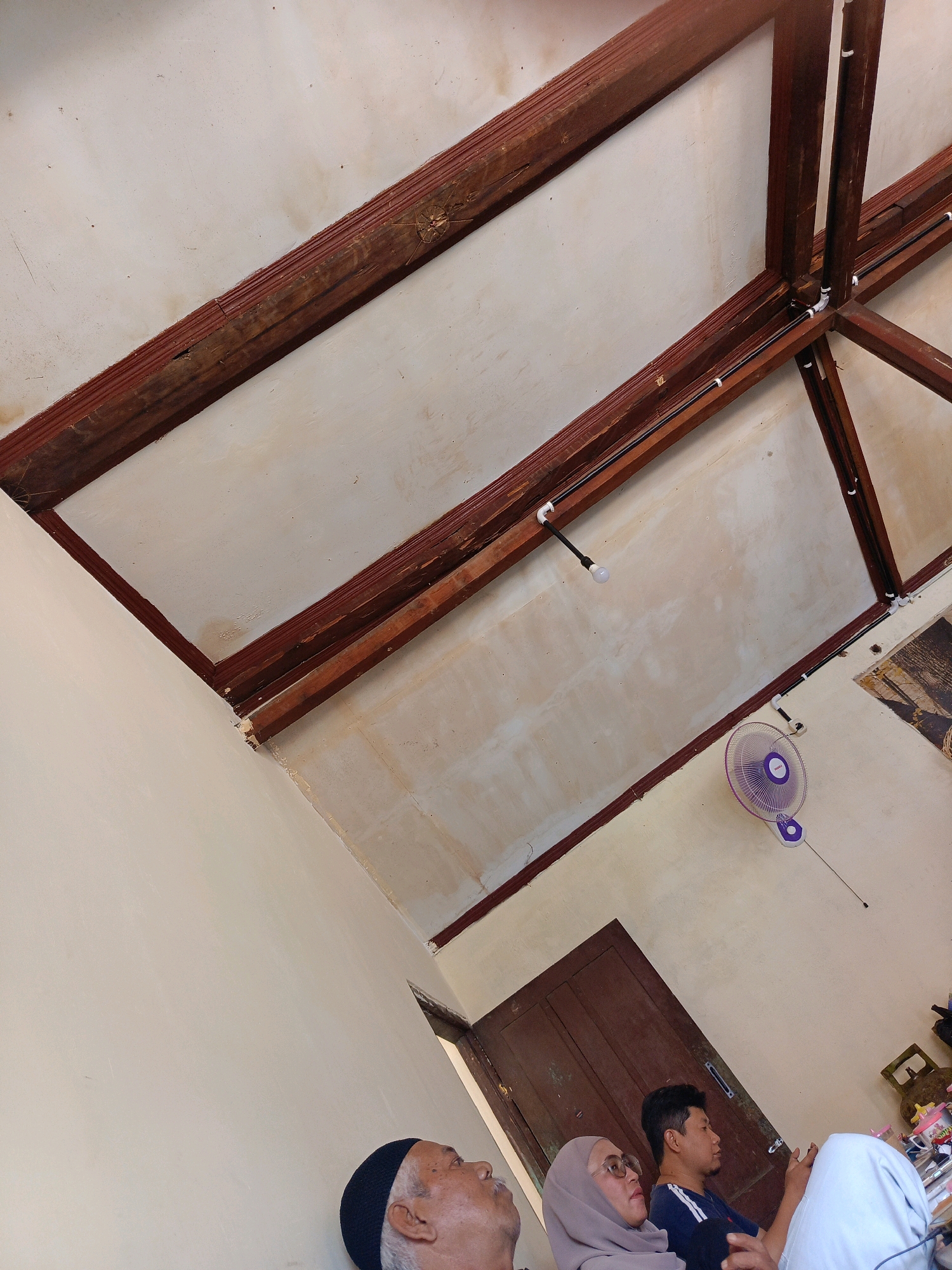 The image shows an interior scene from Kebonagung, Indonesia, with a focus on the wooden ceiling structure and the upper parts of the walls. The ceiling features prominent dark wooden beams that form a geometric pattern against a textured, off-white surface. Electrical conduits and a light fixture are visible along some of the beams. A purple oscillating fan is mounted on the wall near a closed wooden door. In the foreground, several individuals are partially visible, seated and facing to the right, suggesting a gathering or meeting. One person is wearing a black skullcap, another a light gray hijab and glasses, and a third is a young man in a dark blue shirt. The overall impression is of a simple, functional indoor space in a residential or community setting.